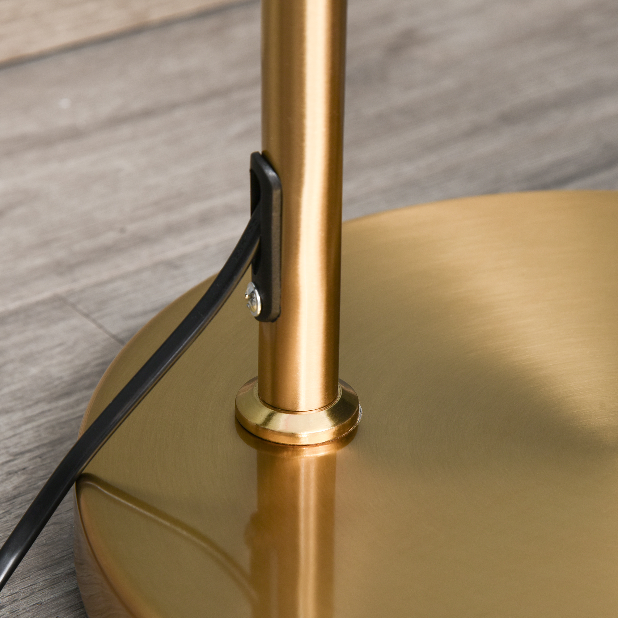 Ein goldfarbener Lampenfuß mit einem schwarzen Kabel. Das Kabel ist mit dem Sockel verbunden. Der Boden ist aus Holz.