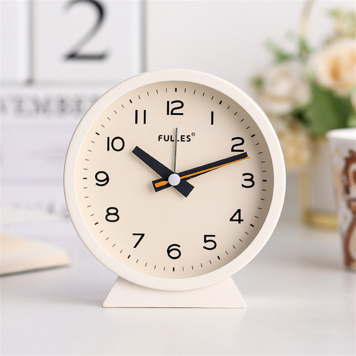 Ein weißer, runder analoger Wecker mit schwarzen und orangenen Zeigern steht auf einer weißen Oberfläche.