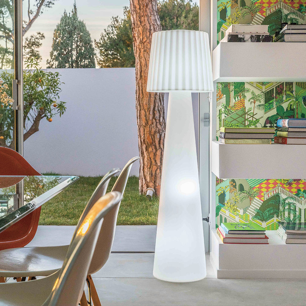 Hohe weiße Stehlampe mit plissiertem Lampenschirm. Sie steht neben einer Wand mit gemusterter Tapete.