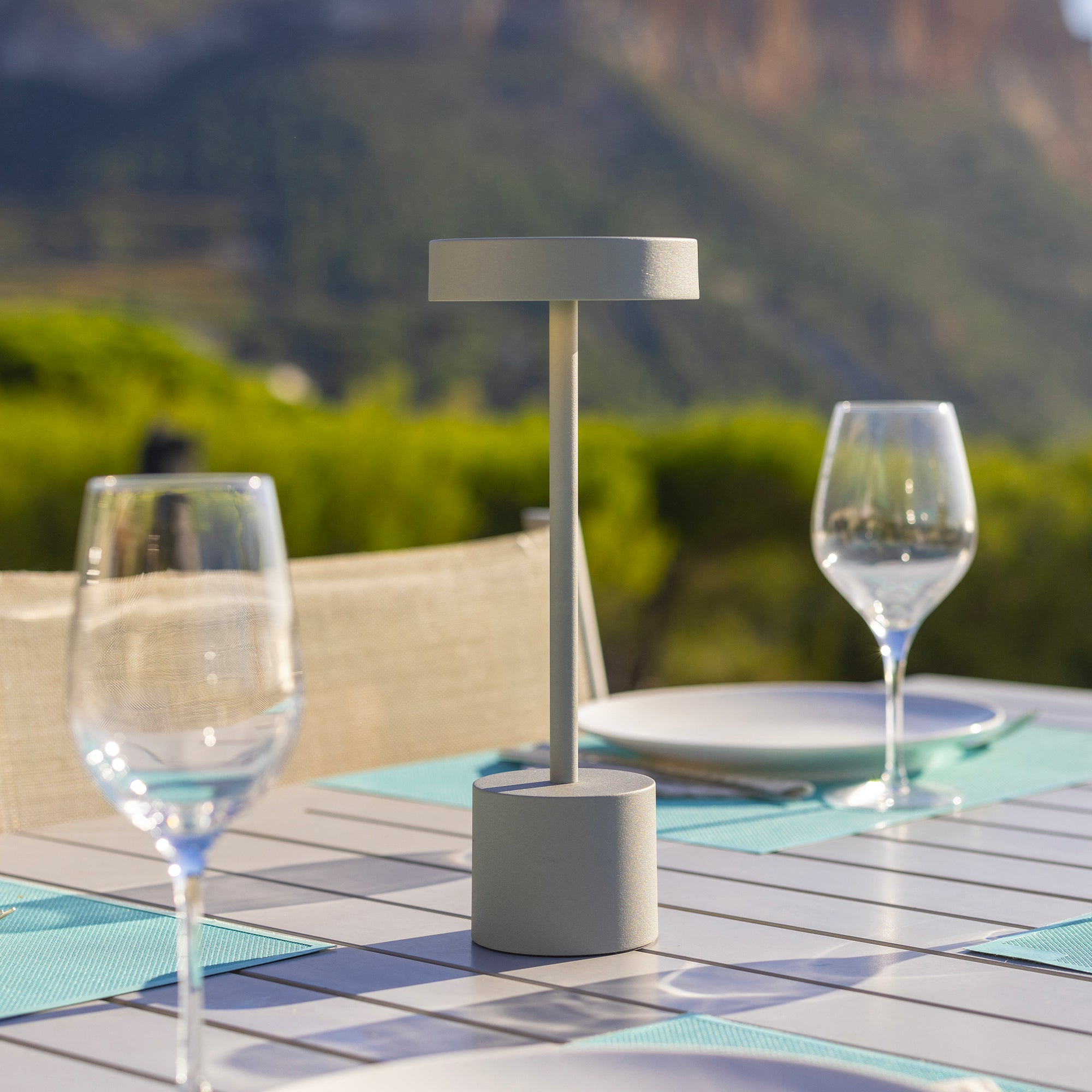 Graue Lampe auf einem Tisch, mit Weingläsern und verschwommenem Gartenhintergrund. Der Tisch ist gedeckt.