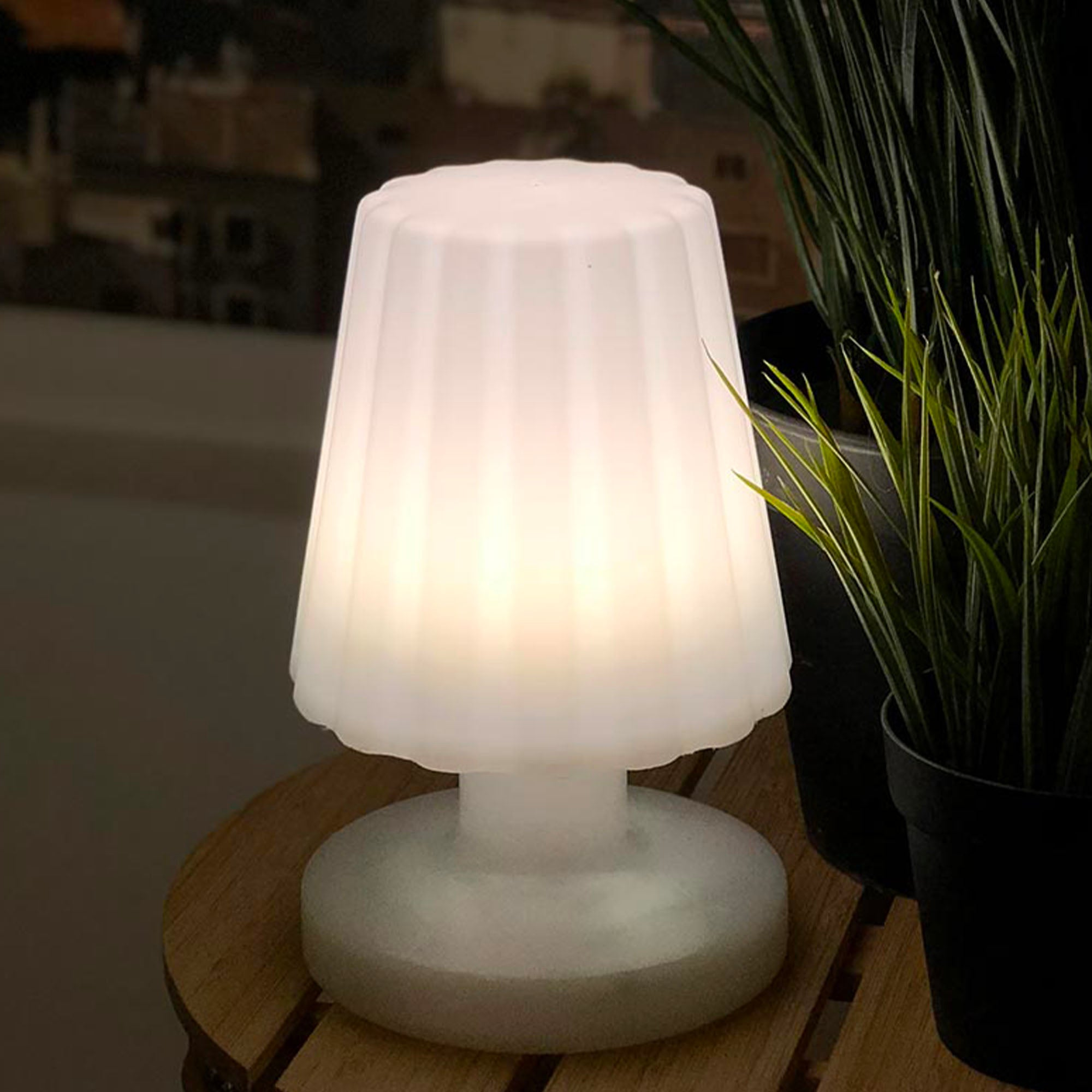 Weiße Lampe mit geriffeltem Lampenschirm auf einem Holztisch. Rechts von der Lampe ist eine Pflanze.