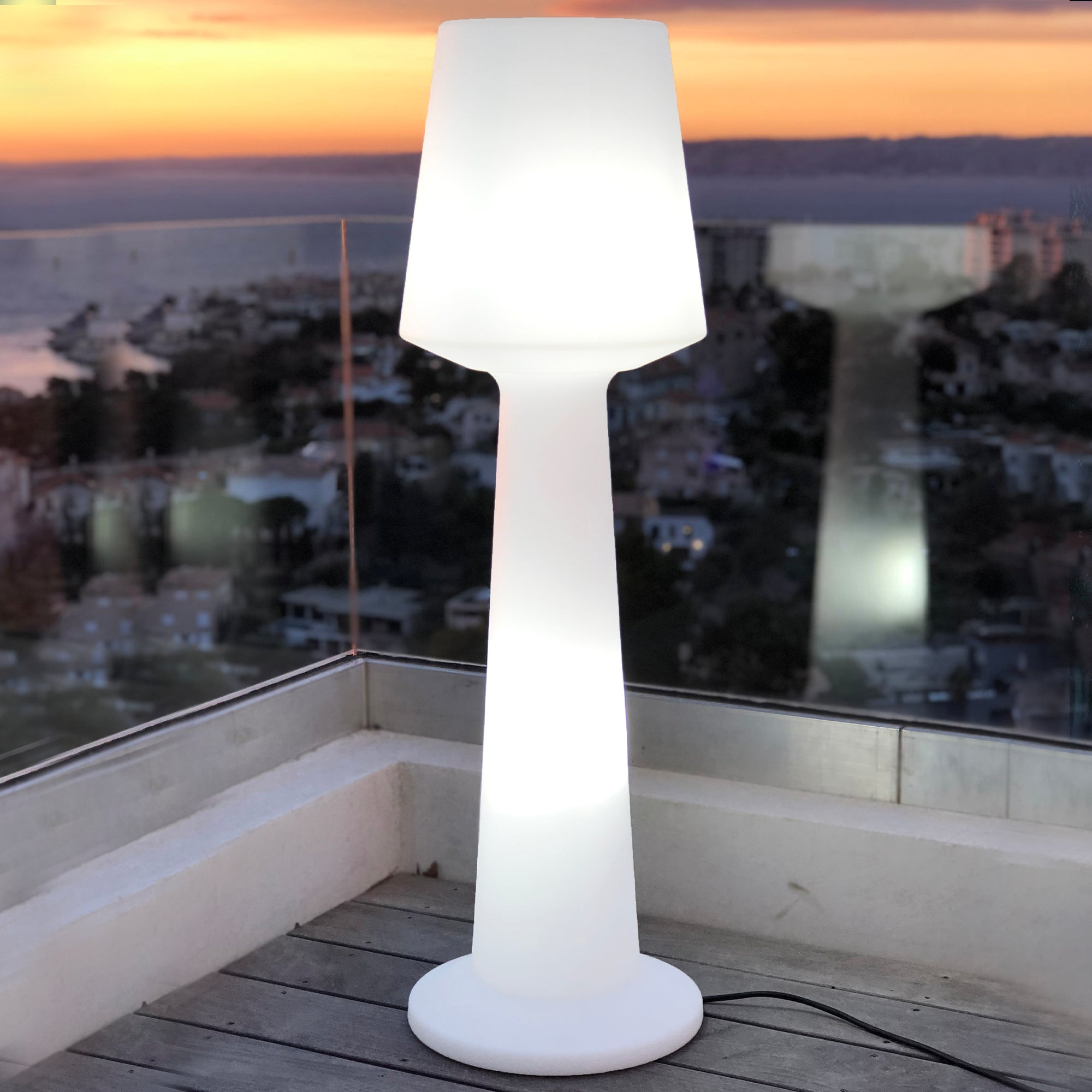 Hohe weiße Außenlampe auf Holzterrasse mit Sonnenuntergang im Hintergrund. Ein Kabel ist sichtbar.