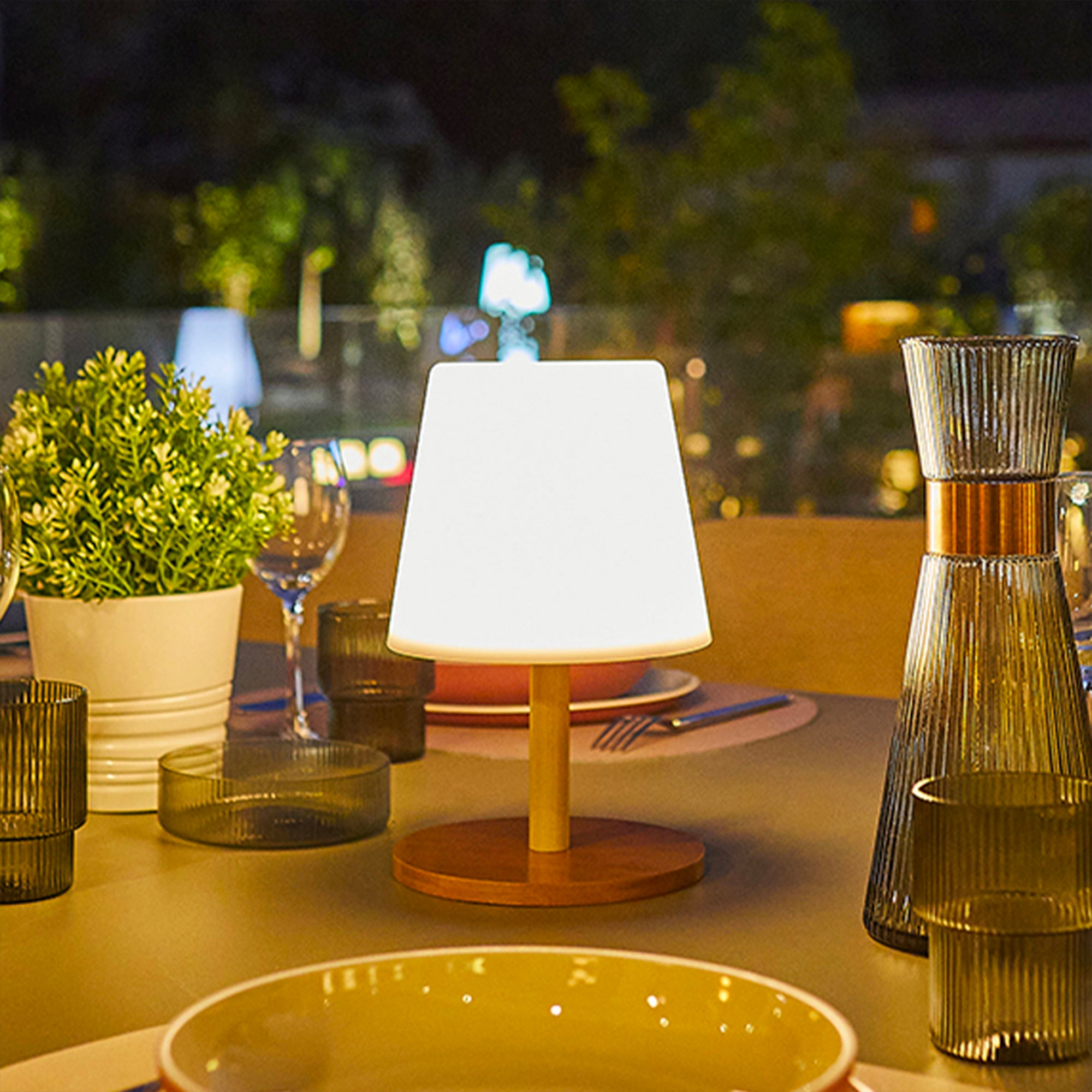 Eine kleine Lampe auf einem Tisch, der zum Essen gedeckt ist. Es gibt Gläser und eine Pflanze.