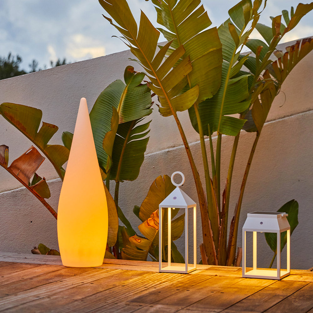 Hohe, weiße Lampe und zwei Laternen auf Holzdeck. Palmenartige Blätter im Hintergrund.