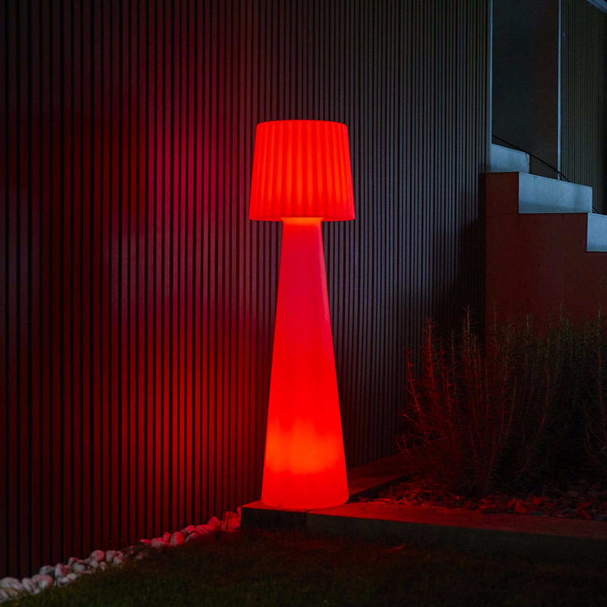 Una lampada rossa alta è illuminata di notte. Ha un paralume e un supporto.