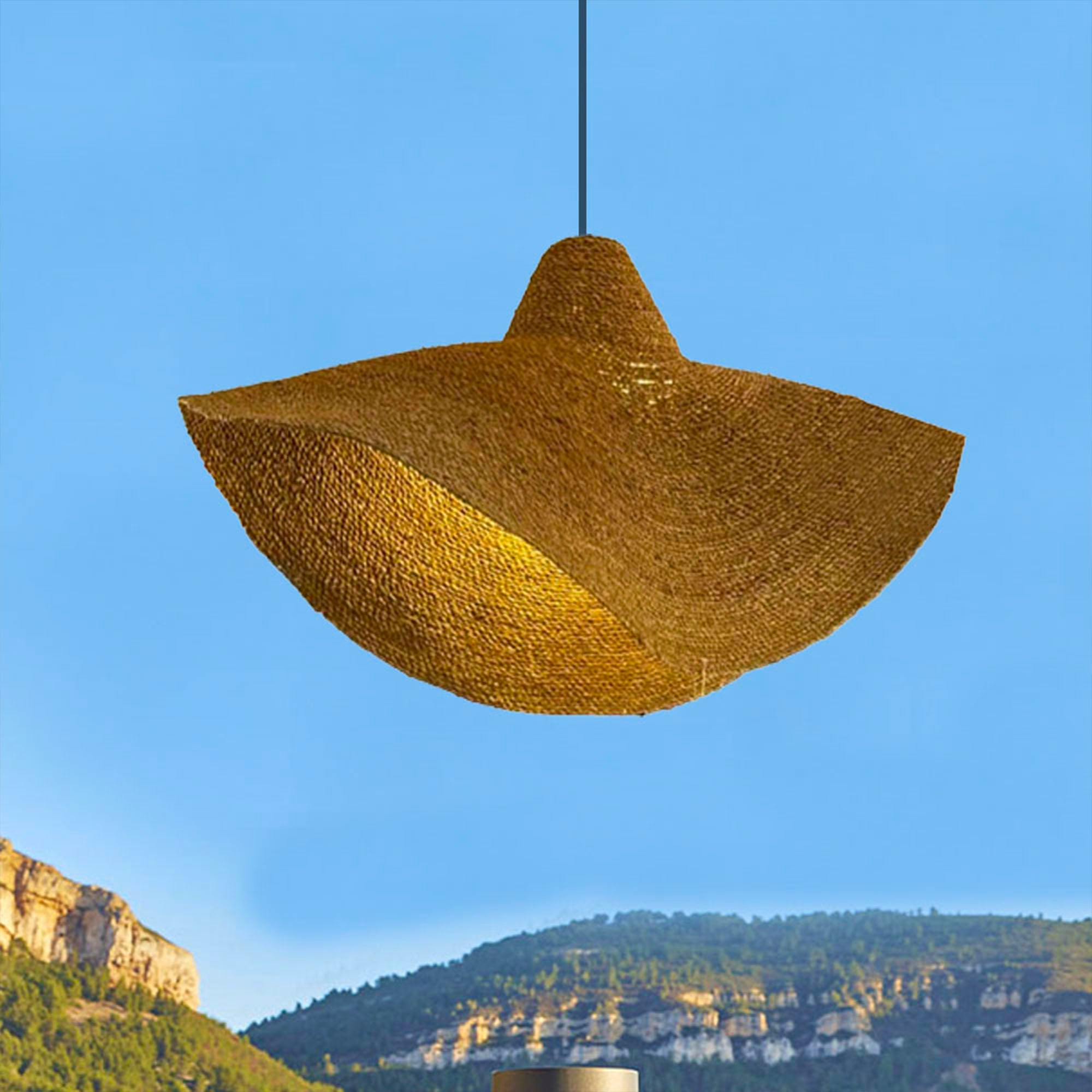 Gewebte, flache, hutartige Pendelleuchte, die vor blauem Himmel und Bergen hängt.