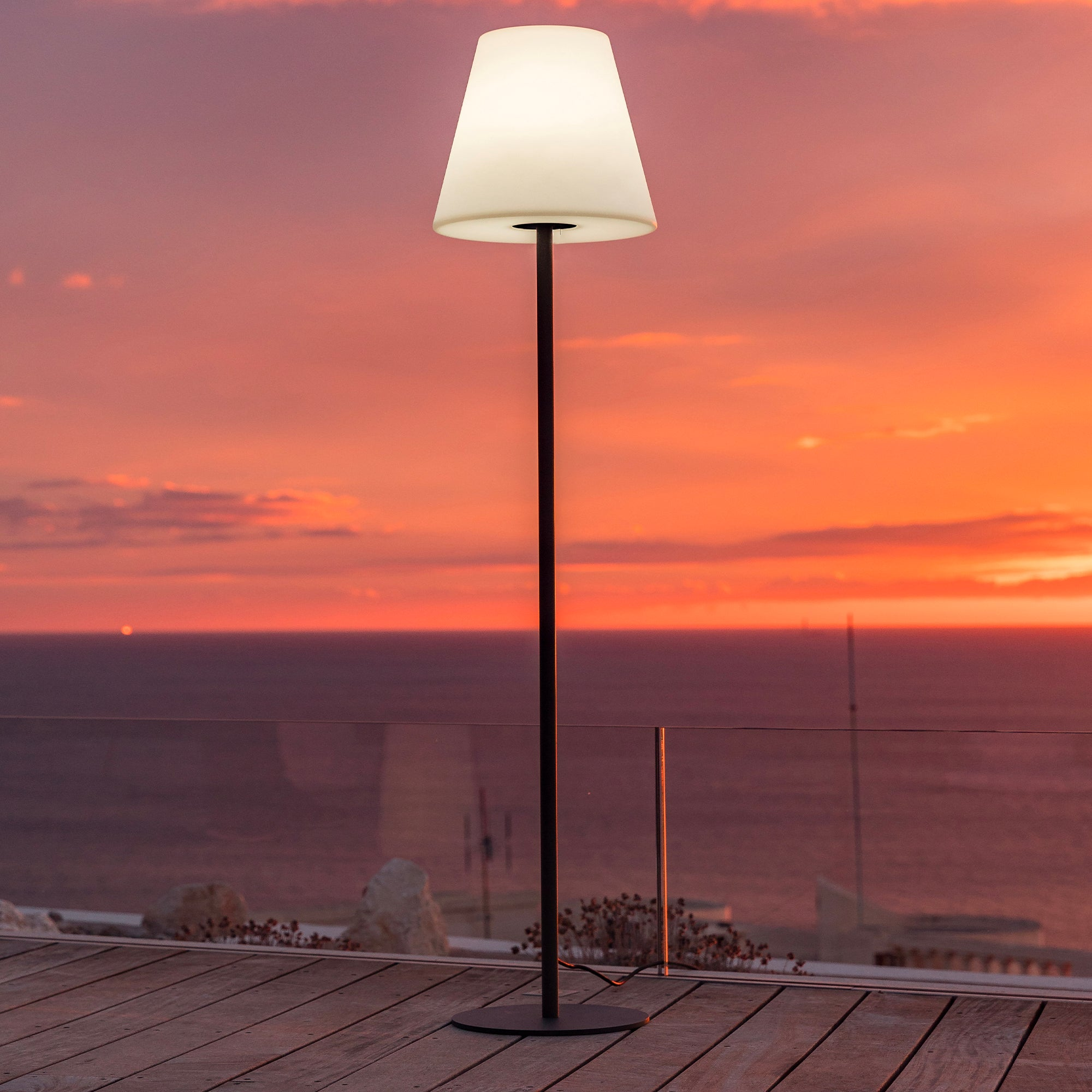 Hohe, weiße Stehlampe mit konischem Schirm auf Holzdeck, Sonnenuntergang im Hintergrund.