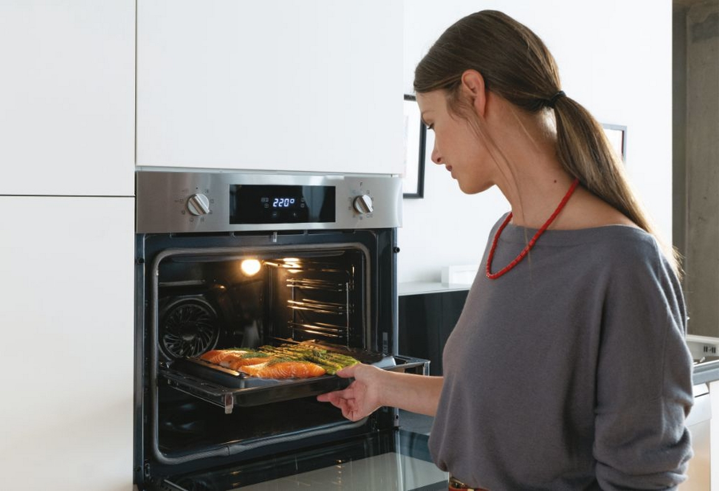 Donna che mette un vassoio di cibo, salmone e asparagi, in un forno a incasso. Il forno è aperto.