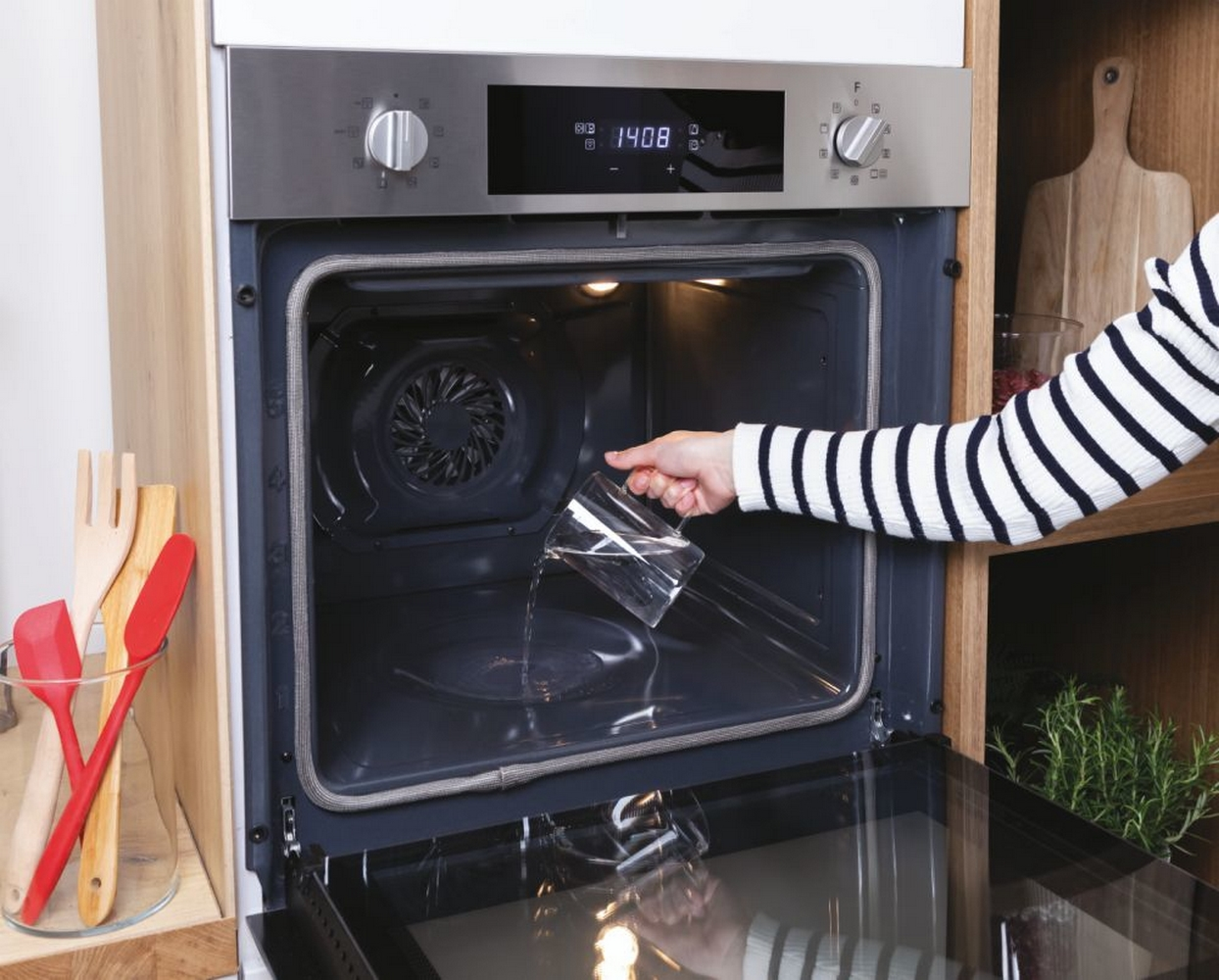 Donna che versa acqua in un forno. Lo sportello del forno è aperto e l'interno è visibile. Utensili di legno vicini.