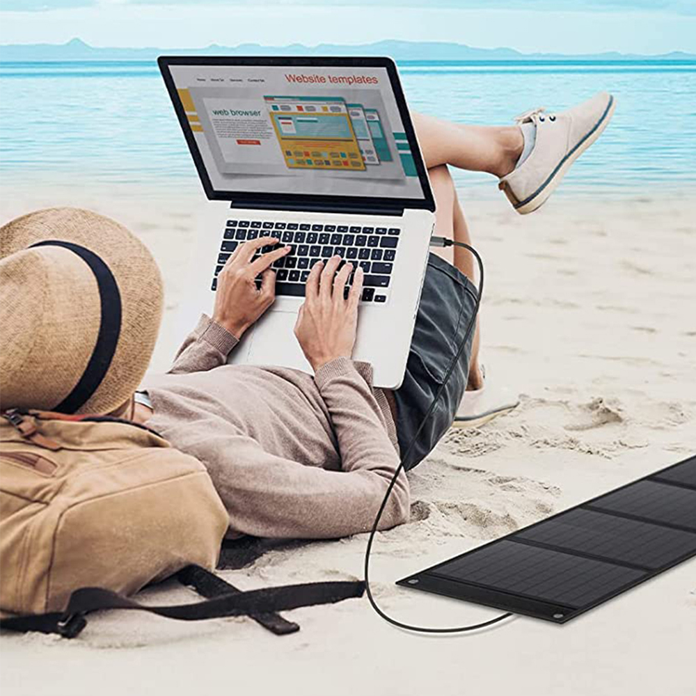 Person am Strand, die einen Laptop benutzt, der an ein Solarpanel angeschlossen ist, mit einem Rucksack in der Nähe.