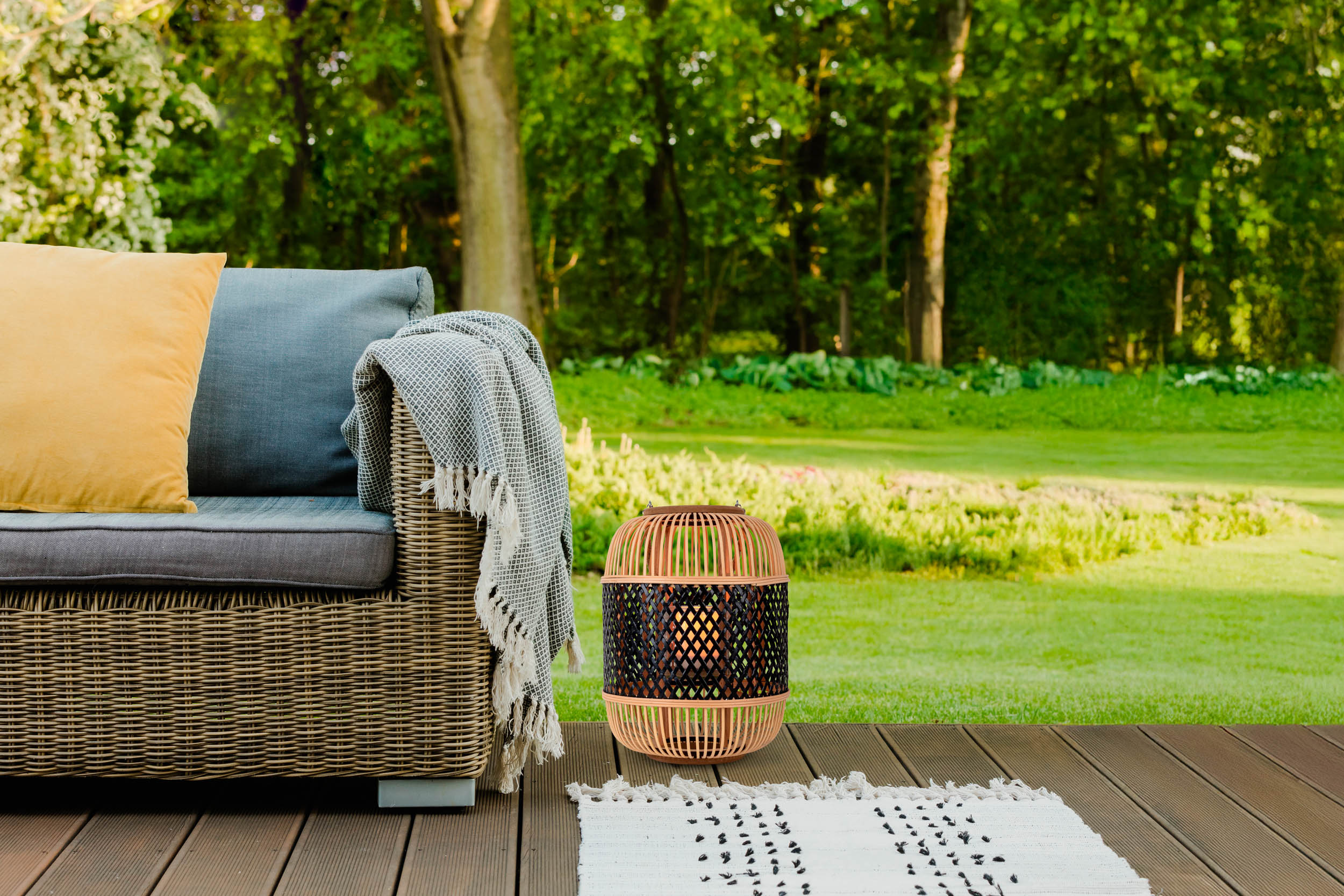 Szene im Freien mit Sofa, Decke und Laterne auf Holzterrasse. Grünes Gras und Bäume im Hintergrund.