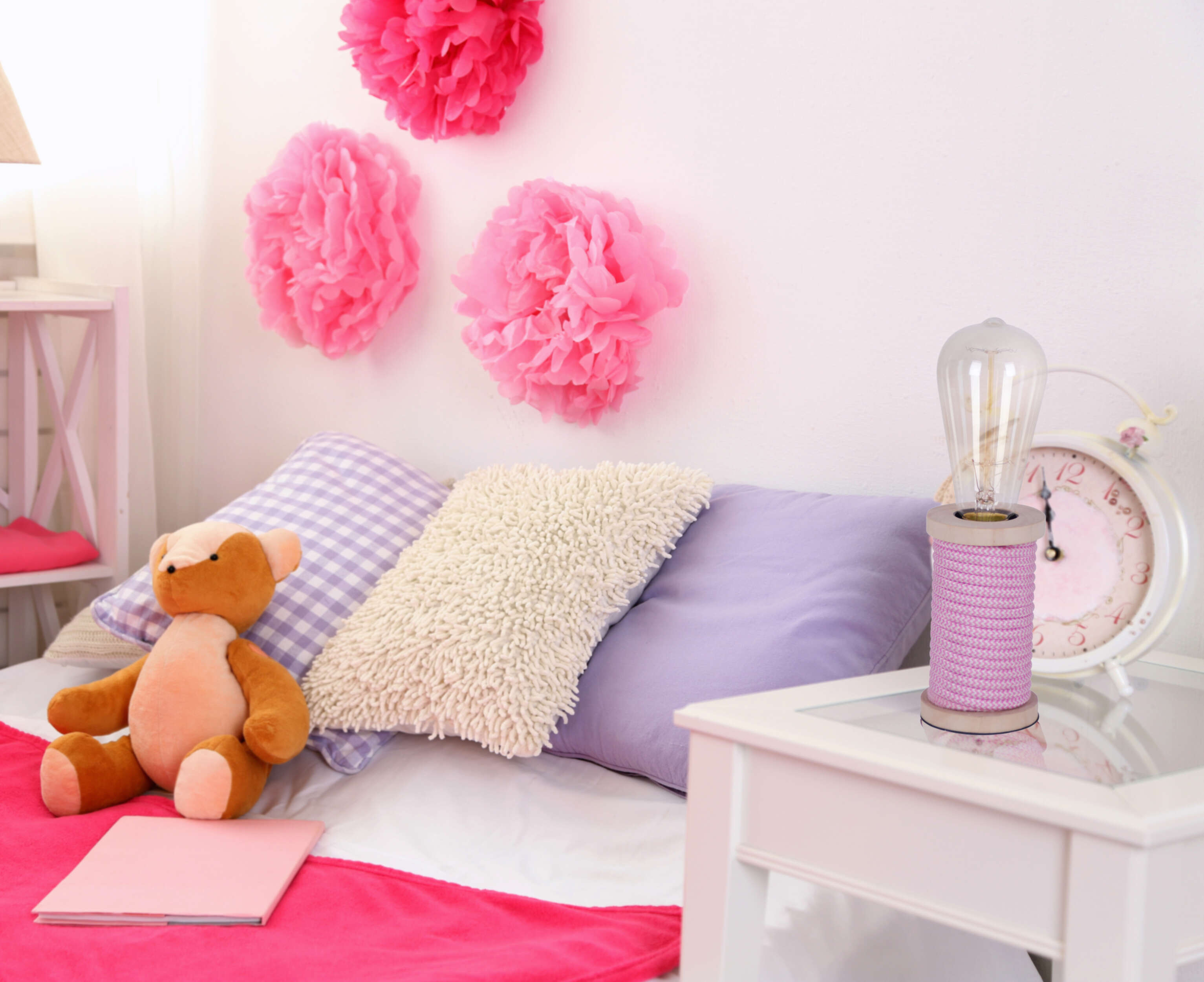 Rosa Deko in einem Raum. Rosa Pompons, ein Bett mit Kissen, eine Lampe, eine Uhr und ein Teddybär.