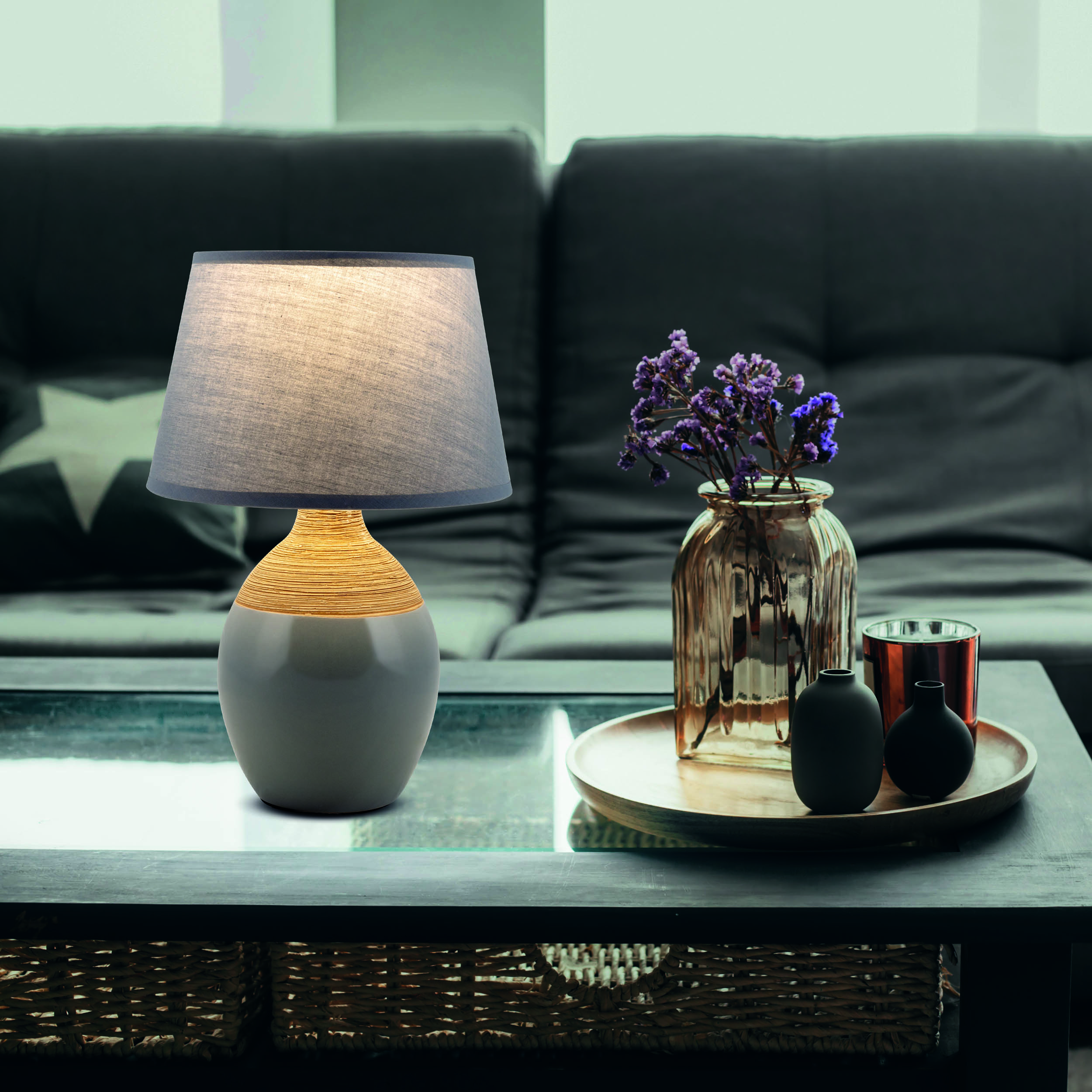 Tischlampe auf einem dunklen Tisch. Die Lampe hat einen grauen Schirm und einen beigen, umwickelten Sockel. Hintergrund: Sofa, Vase, Deko.