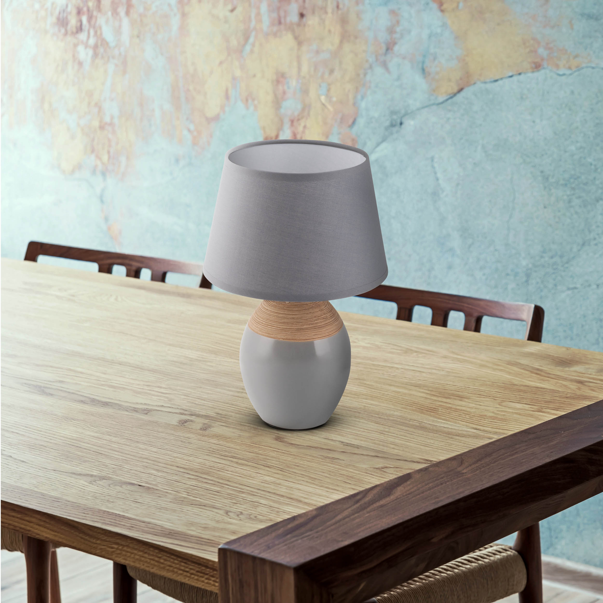 Eine graue Lampe mit grauem Lampenschirm steht auf einem Holztisch. Im Hintergrund eine rustikale Wand.