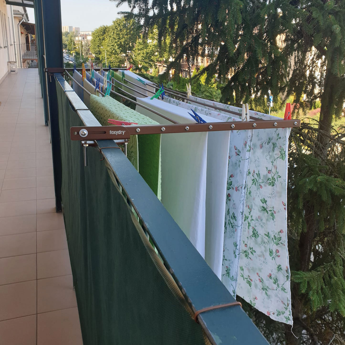 Balkon mit Wäscheständer mit Kleidung. Grüne und weiße Stoffe hängen. Bäume und Stadt im Hintergrund.