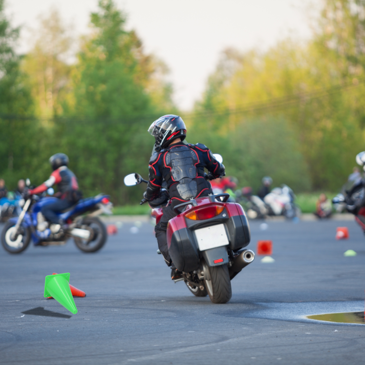 Motorcycles on a track. One rider is in front. Green cones are visible. Trees in the blurred background.