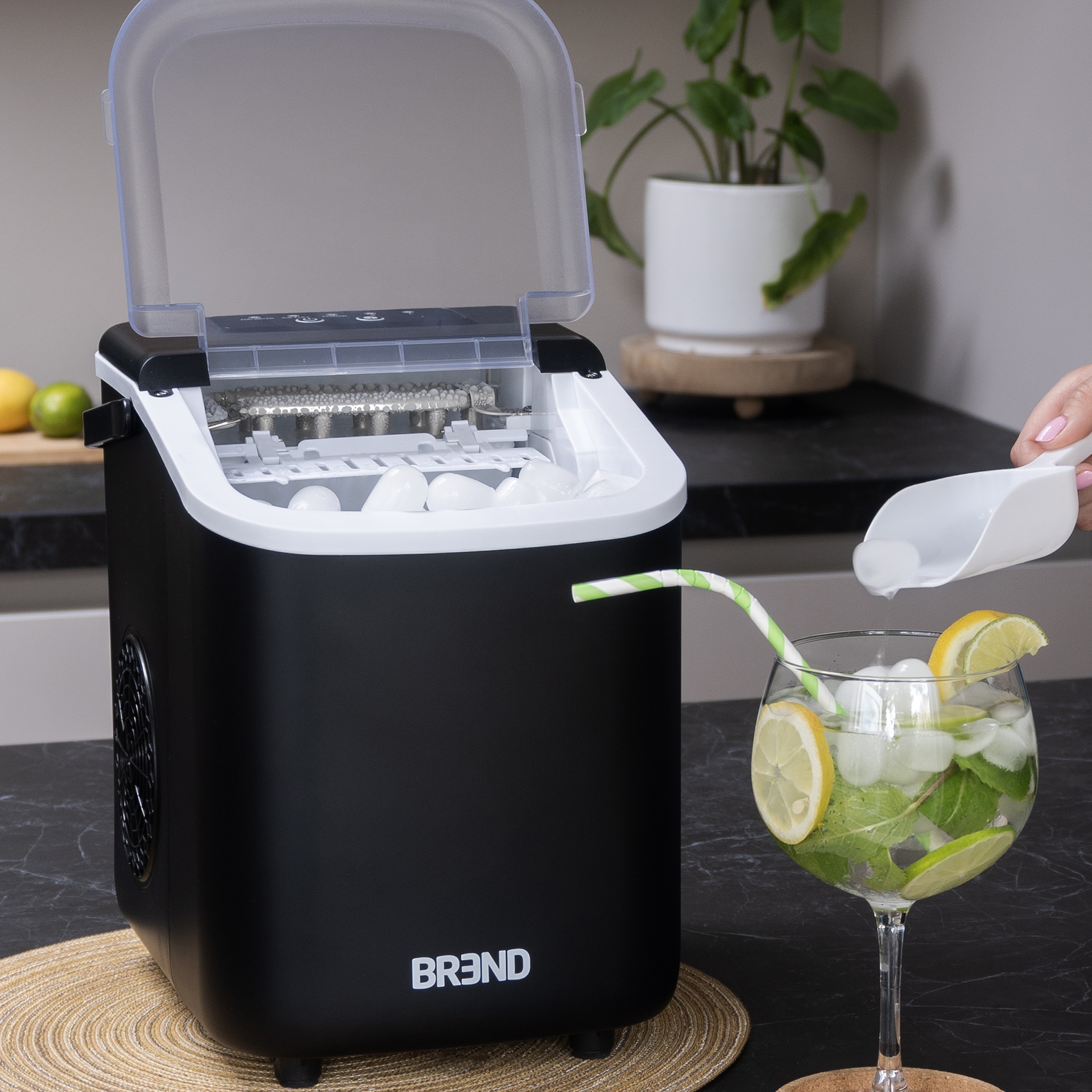 Black ice maker with open lid, ice cubes inside. A glass of cocktail with a straw is on the right.