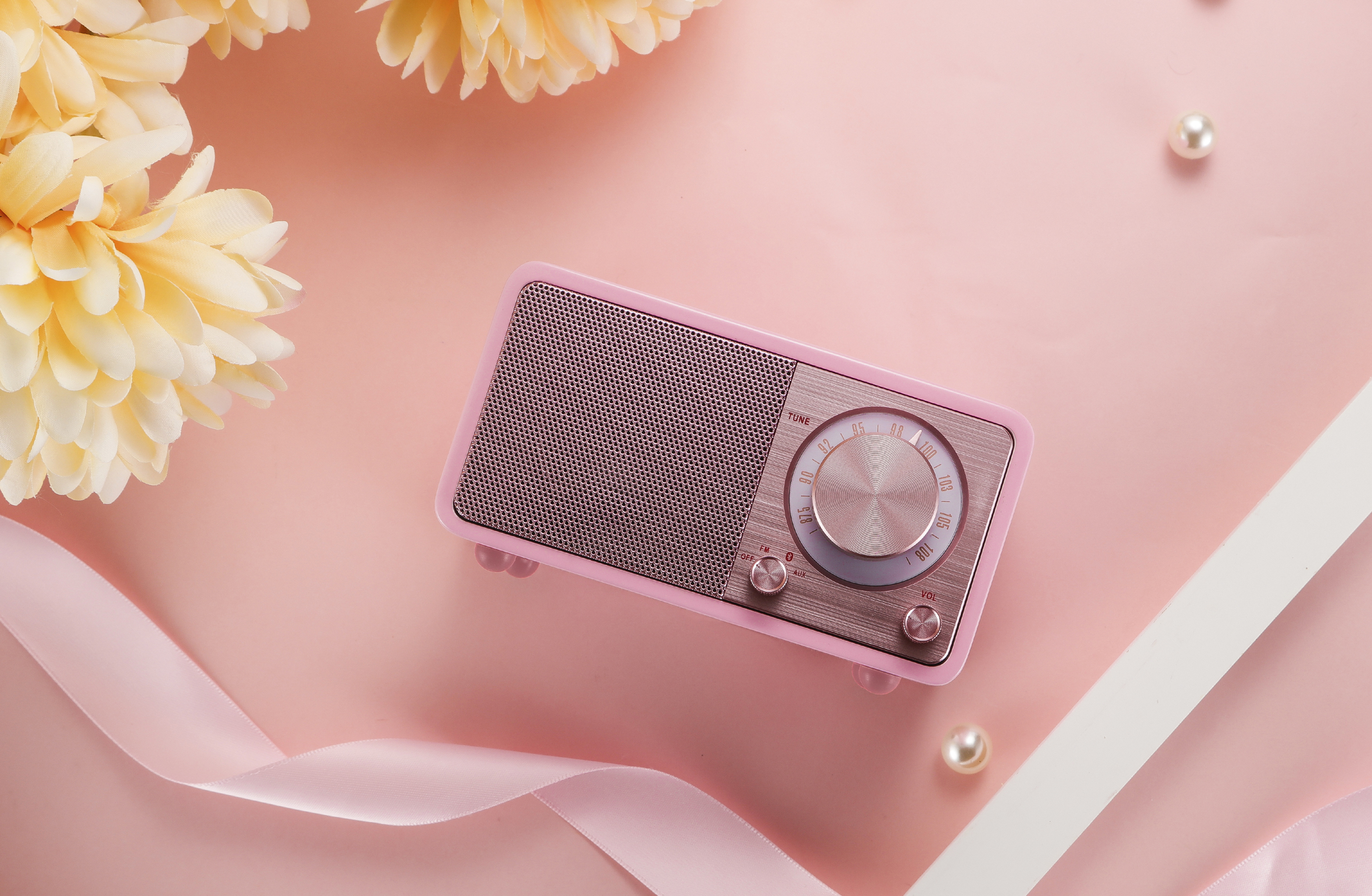 Różowe radio retro na różowej powierzchni, z żółtymi kwiatami i białą wstążką w tle, z perłami.