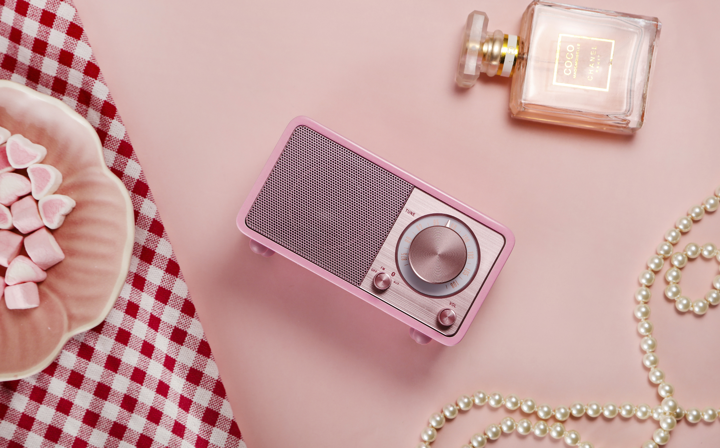 Różowe radio na różowej powierzchni, z talerzem pianek, perfumami Chanel i perłami, na różowym tle.