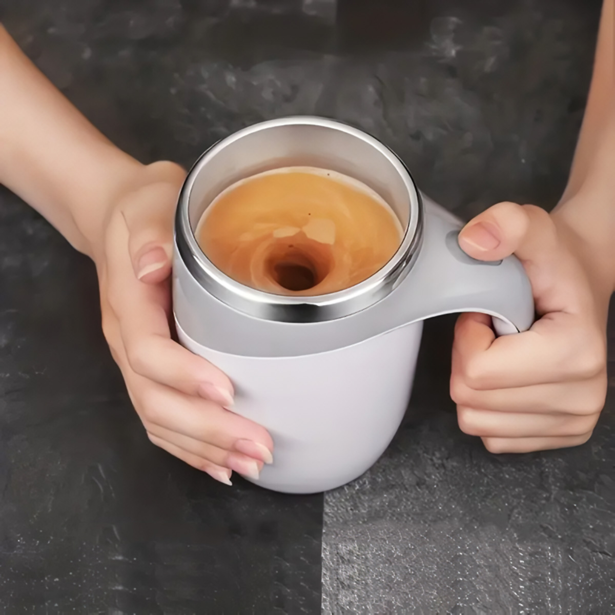 Manos sosteniendo una taza de café blanca y plateada con líquido arremolinado dentro. El fondo está borroso.