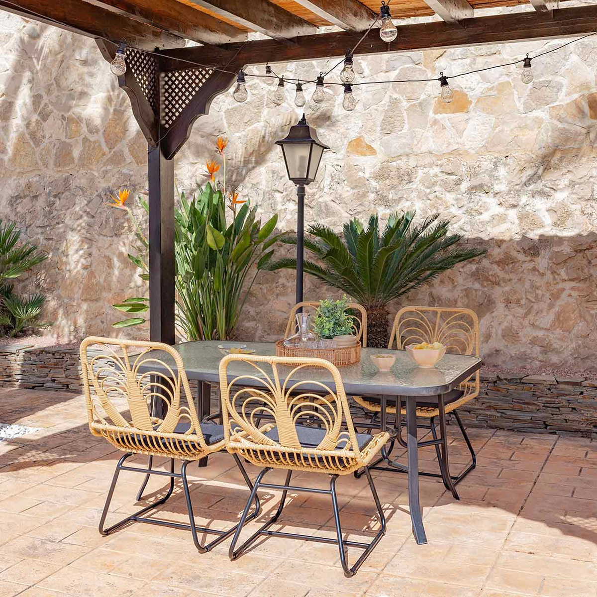 Juego de comedor al aire libre con mesa, sillas y luces de cadena. Fondo de pared de piedra.