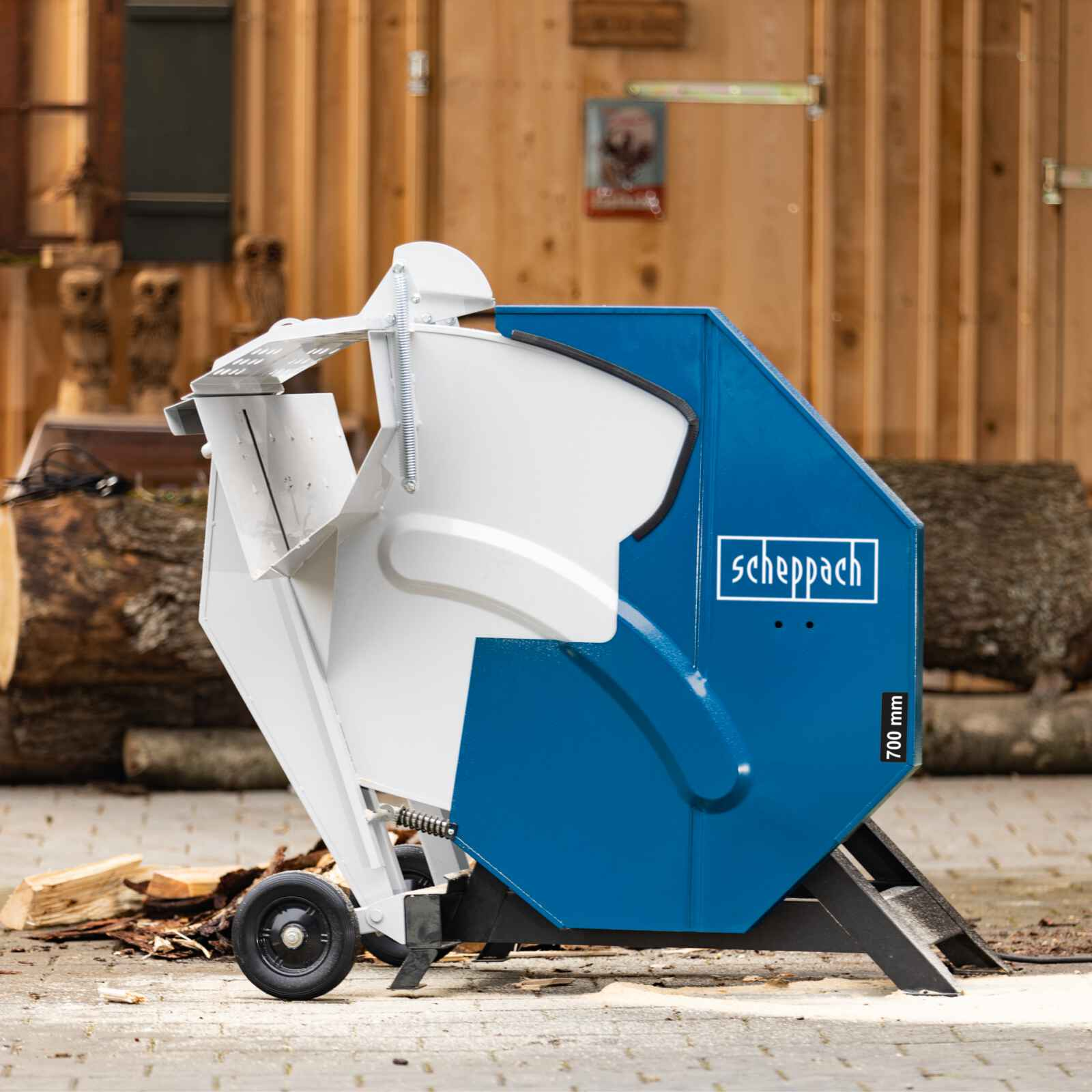 Scheppach houtzaag, blauw en wit. Een gezaagde stam ligt op de machine. Buiten met hout op de achtergrond.