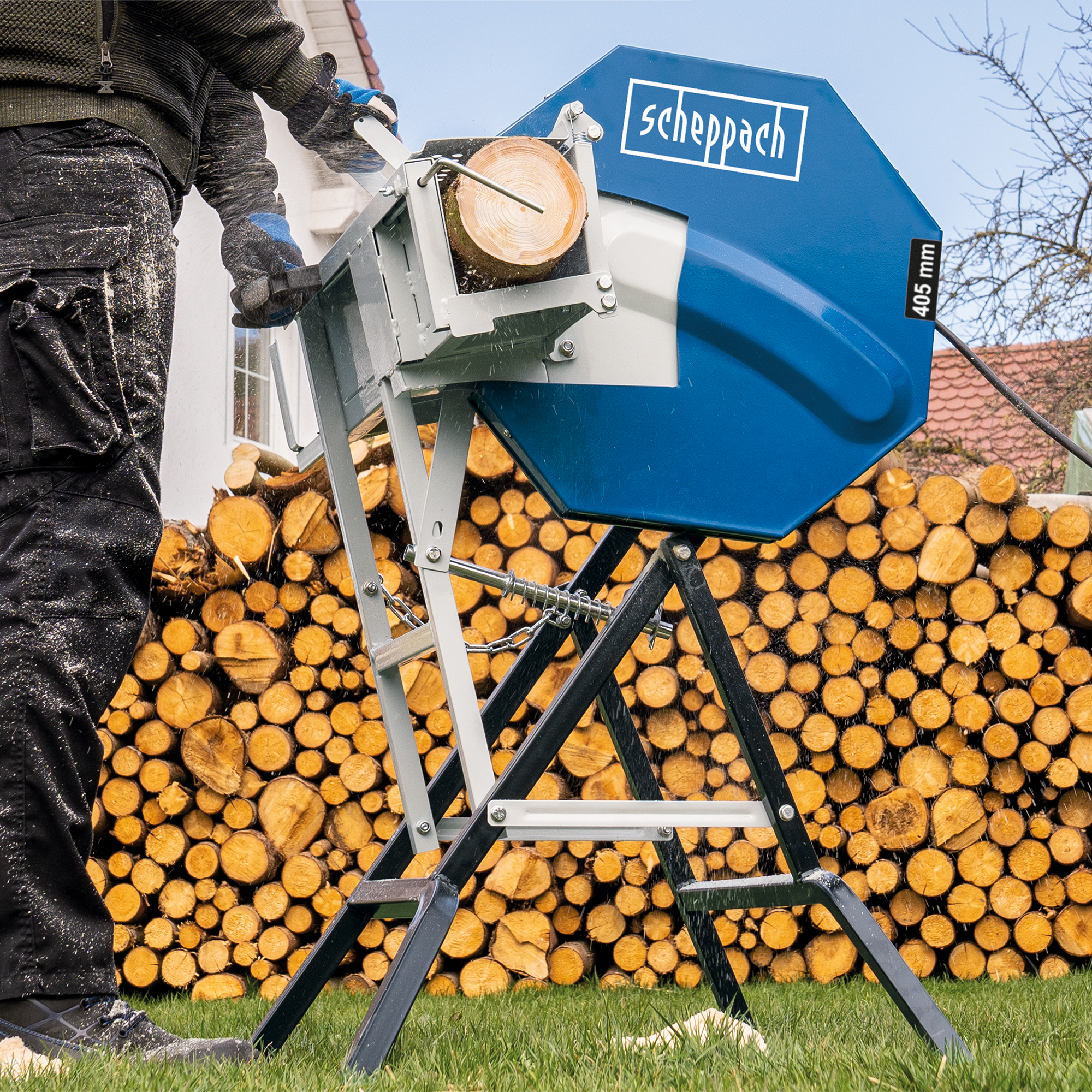 Man zaagt een stam met een Scheppach zaag. Hout opgestapeld op de achtergrond. Groen gras en zonlicht.