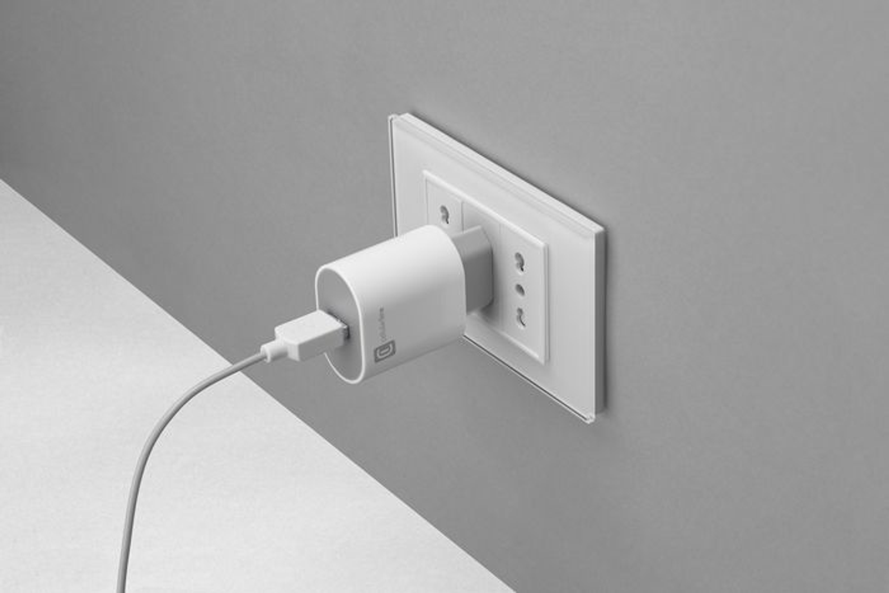 Cargador blanco enchufado a una toma de corriente, contra una pared gris.