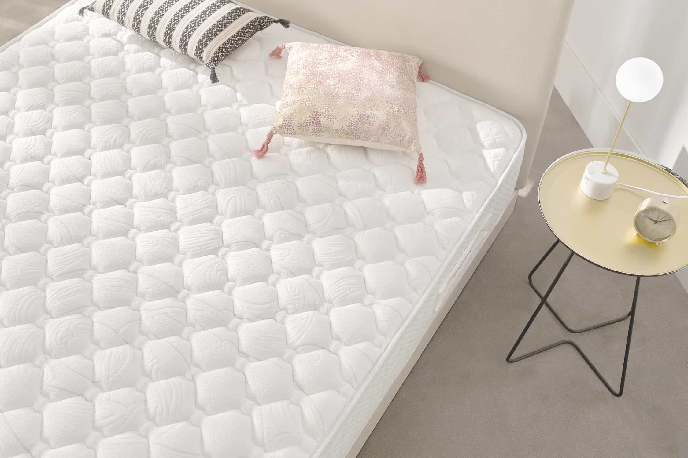 Colchón blanco en marco de cama. Almohadas y lámpara en mesita. Habitación luminosa.