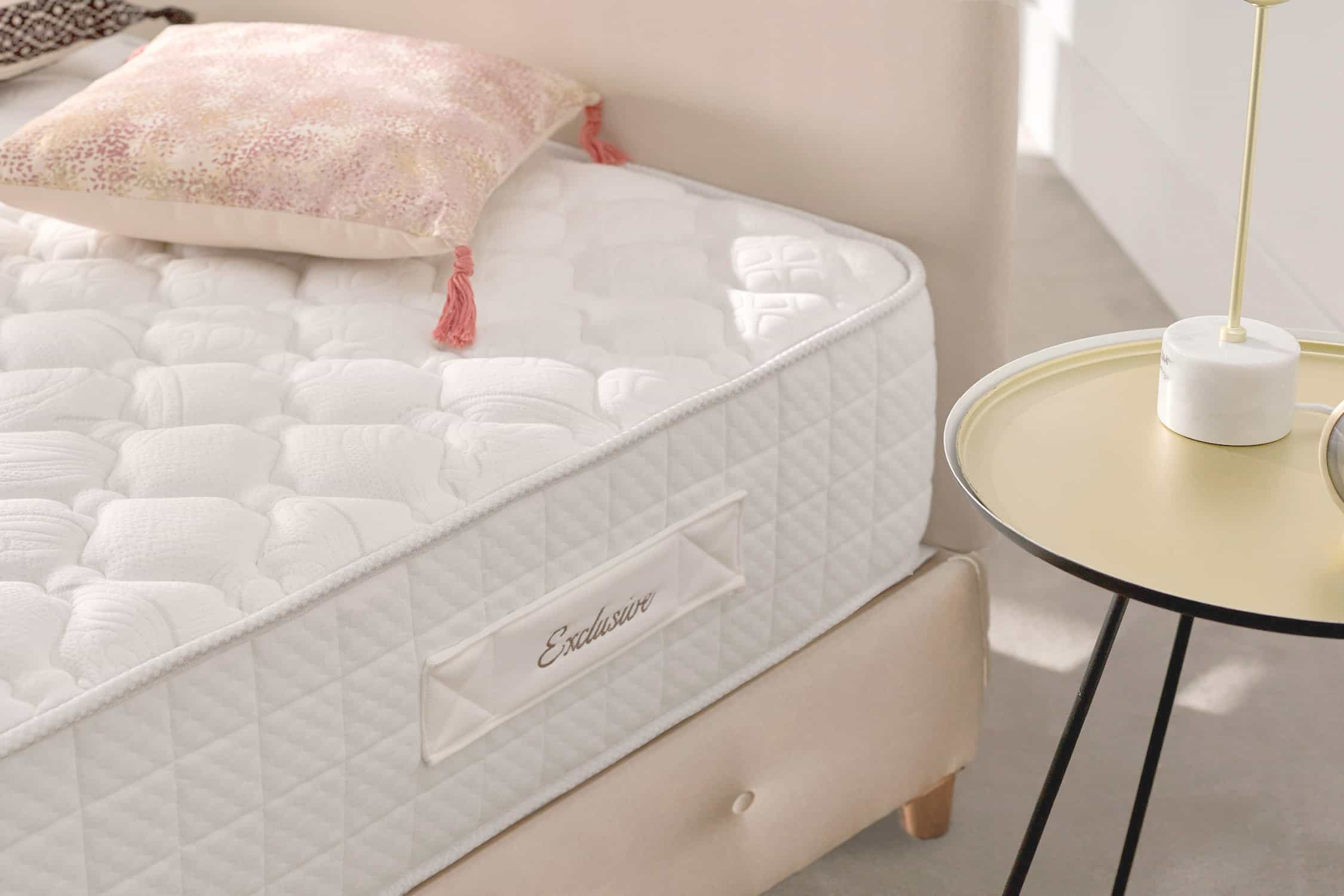 Una cama con colchón blanco en un dormitorio, con una mesita y almohada.