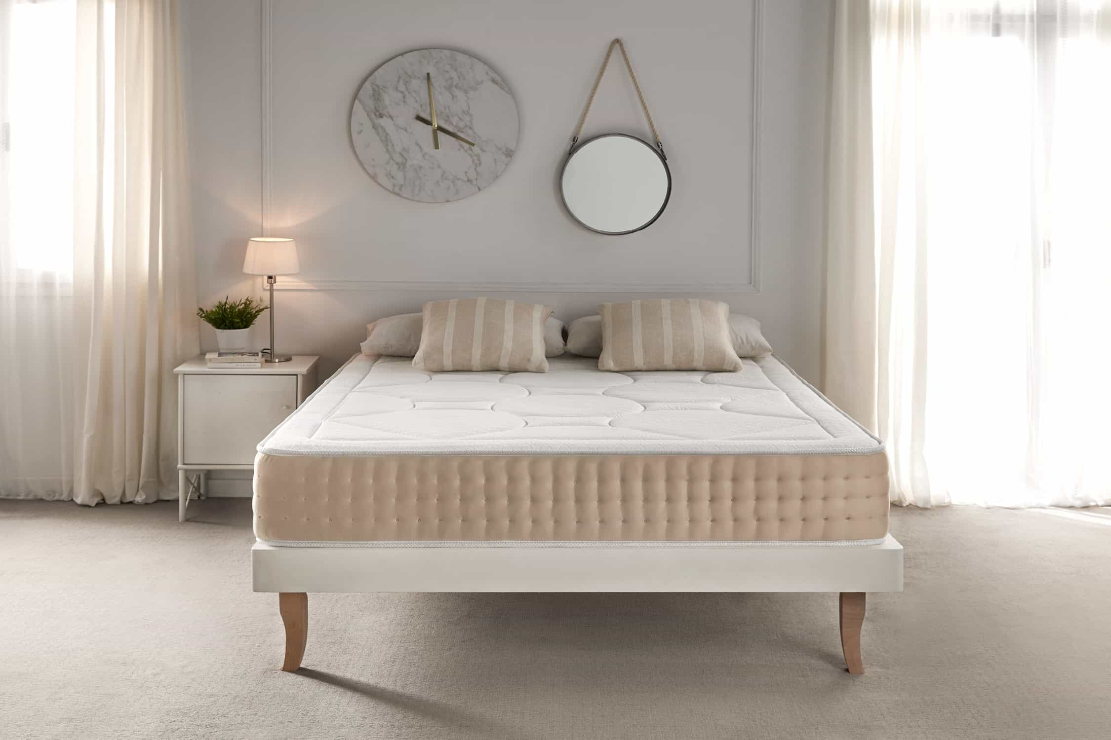 Escena de dormitorio con cama, colchón, almohadas, mesita de noche con lámpara, espejo, reloj y cortinas.