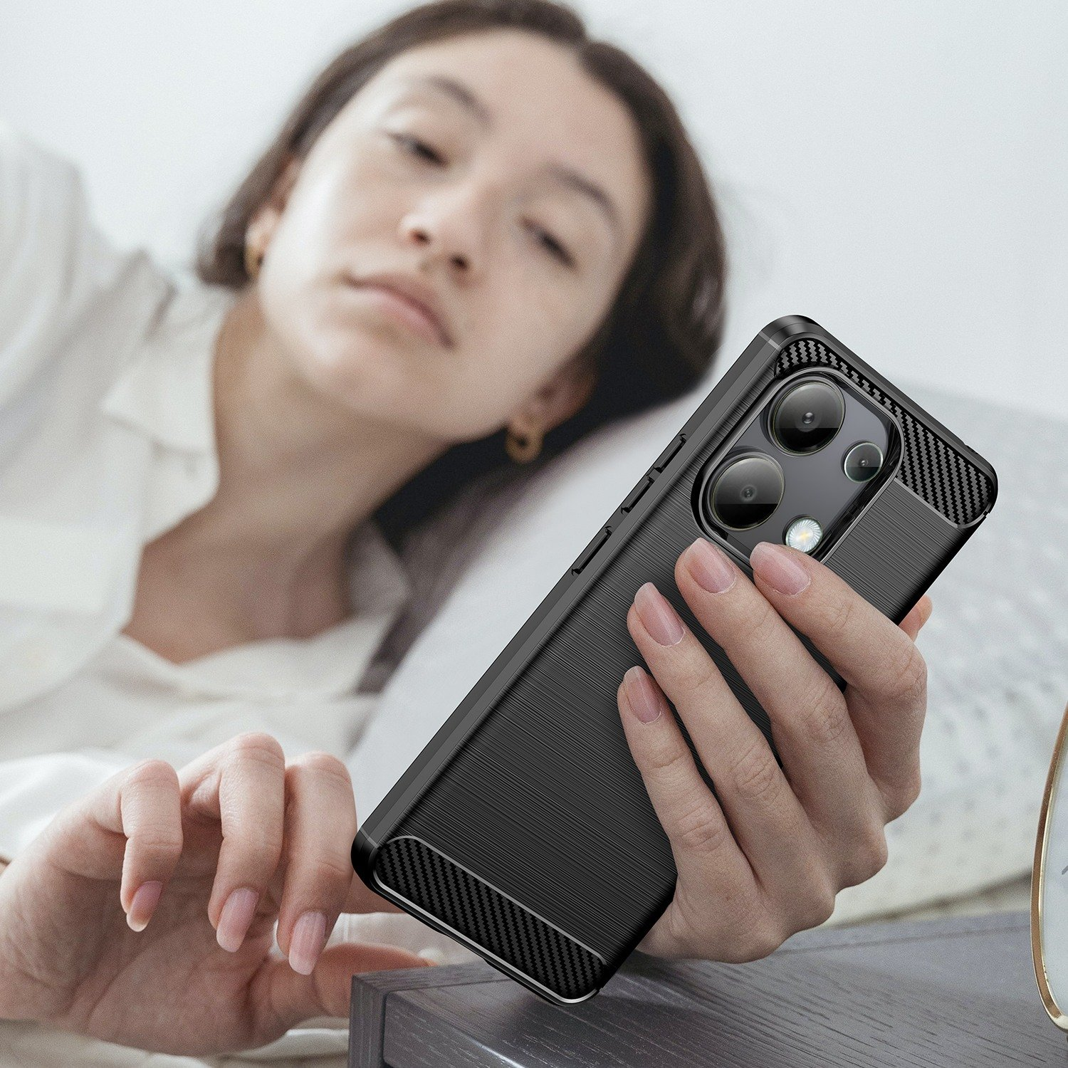 Una mujer sosteniendo un teléfono negro con funda. El teléfono está sobre una almohada. La funda tiene textura de fibra de carbono.