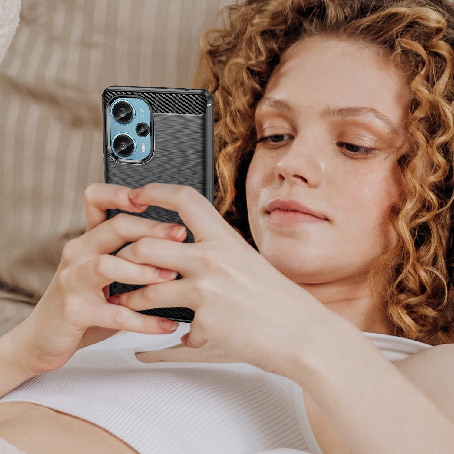 Mujer con cabello rizado mirando un teléfono con funda negra. Camiseta sin mangas blanca. Fondo beige.