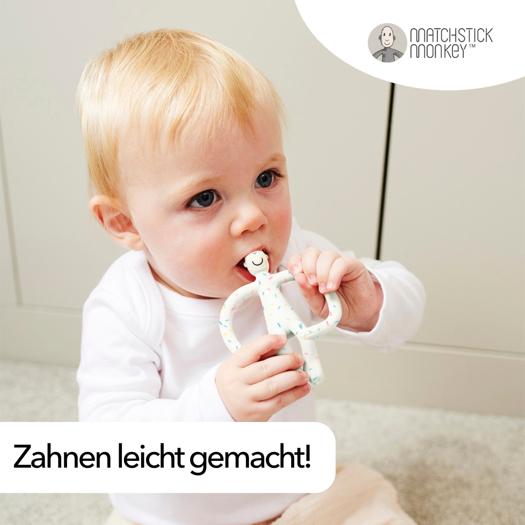 Ein Baby hält und kaut auf einem weißen und bunten Beißring. Der Markenname ist Matchstick Monkey.