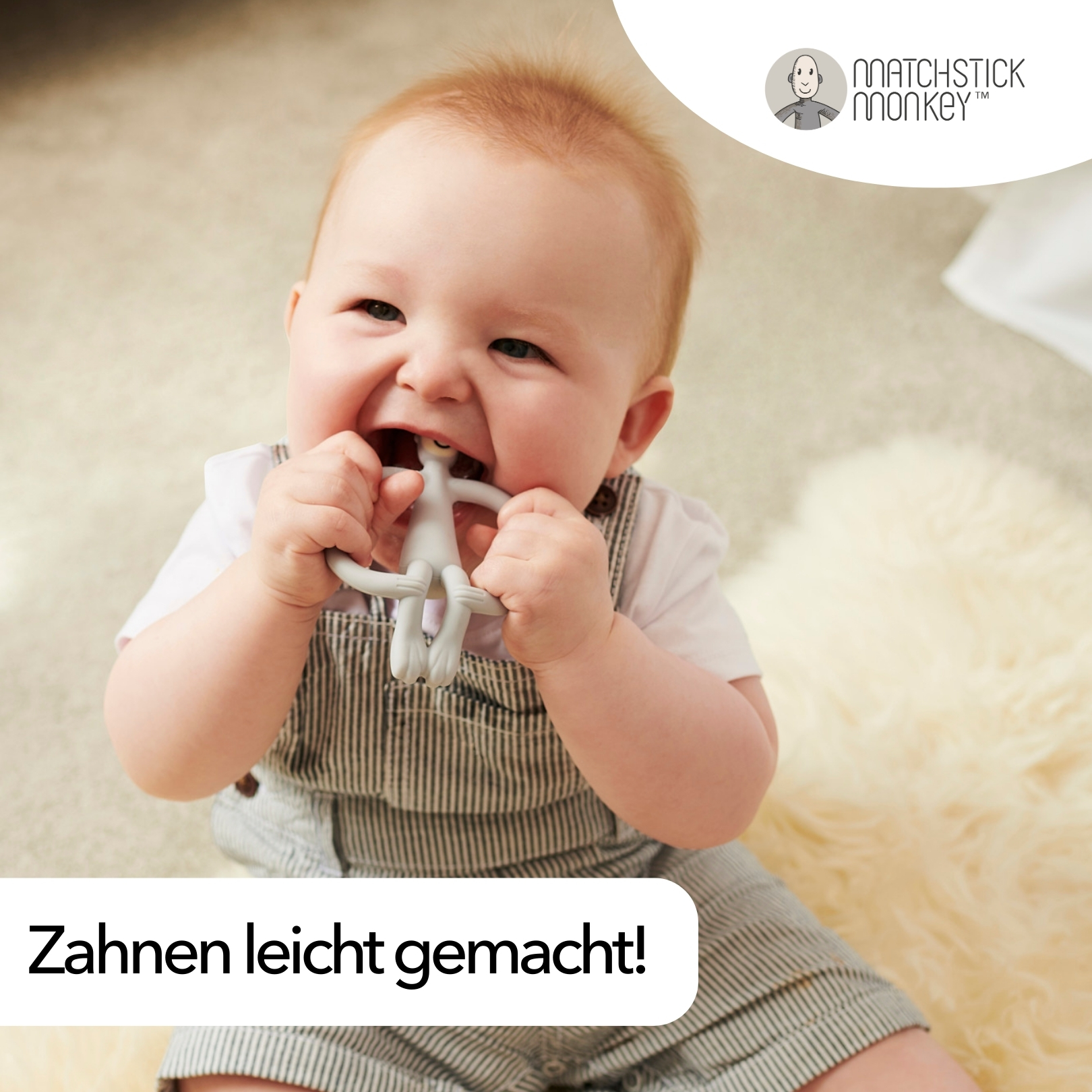 Ein Baby lächelt und kaut auf einem grauen, affenförmigen Beißring. Das Baby trägt gestreifte Latzhosen.