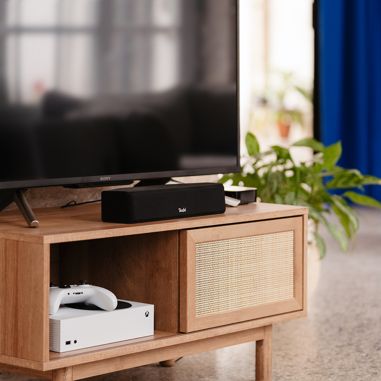 Zwarte luidspreker op een houten kast met een tv erboven. Een spelconsole en een controller staan op de kast.
