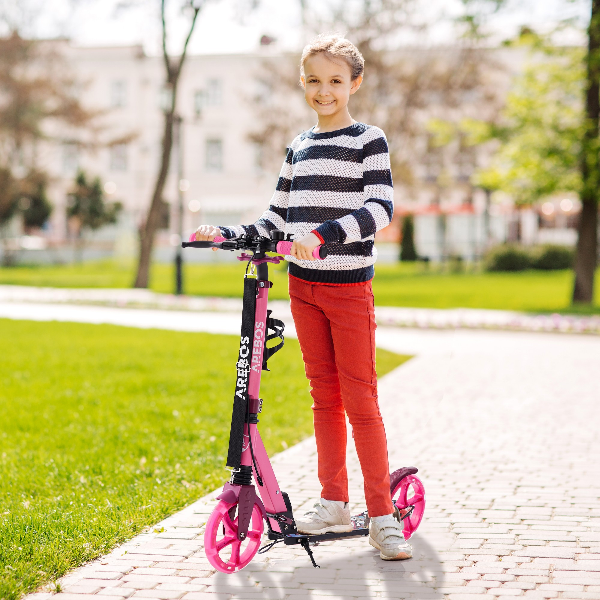 Ein junges Mädchen mit einem Roller in einem Park. Sie lächelt und schaut in die Kamera.