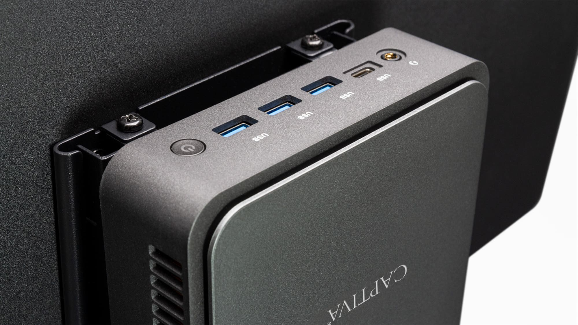 Ciemnoszary komputer przymocowany do czarnego uchwytu monitora, z portami USB i przyciskiem zasilania.