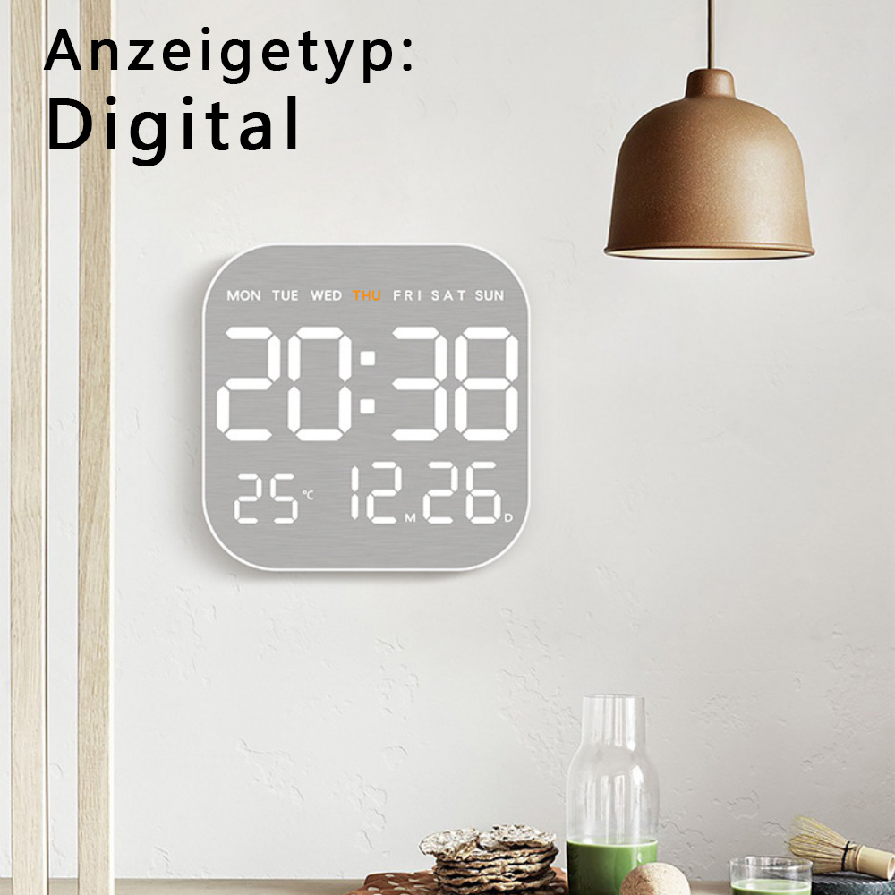 Digitaluhr zeigt Zeit und Datum. Die Uhr befindet sich an einer weißen Wand, mit Lampe im Hintergrund.