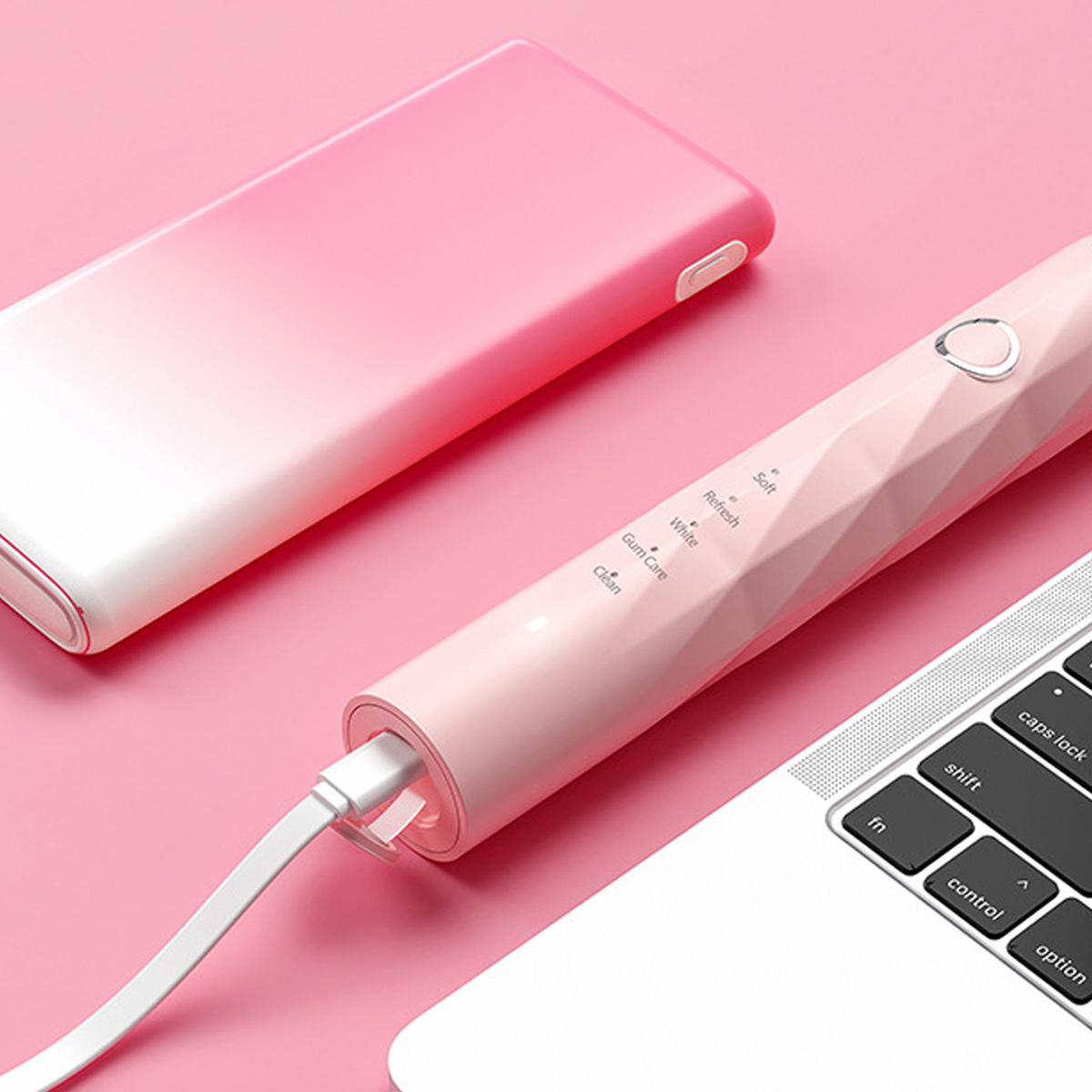 Rosa elektrische Zahnbürste, weißes Kabel in rosa Powerbank, auf rosa Oberfläche. Weißer Laptop teilweise sichtbar.