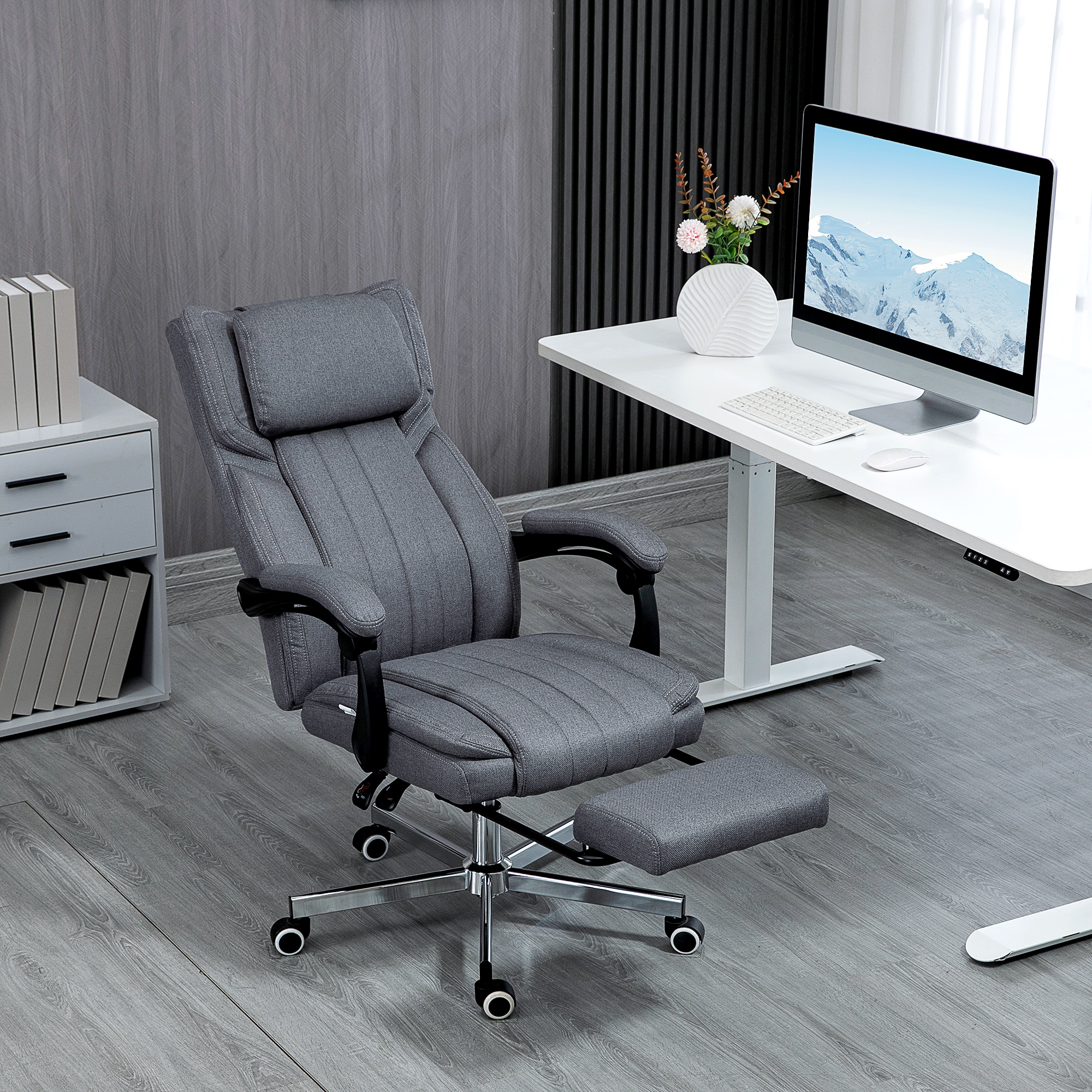 Silla de oficina gris. Reposabrazos y base negros. Escritorio blanco, monitor de computadora y libros en el fondo.