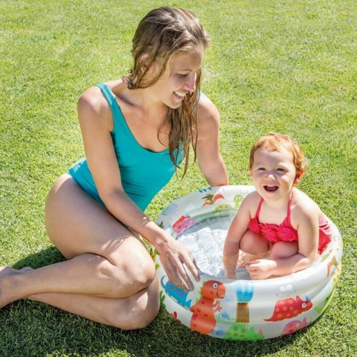 Mujer y niño en una piscina pequeña sobre hierba. La mujer lleva un traje de baño turquesa. Niño traje de baño rosa.