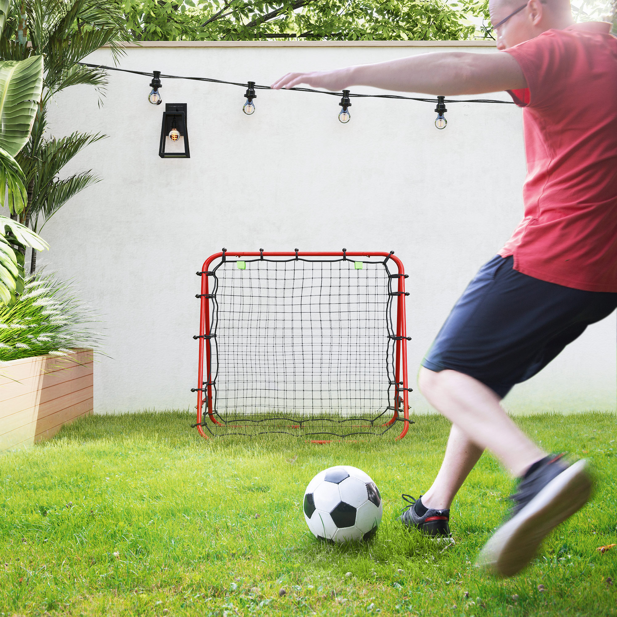 Eine Person schießt einen Fußball in Richtung eines rot-schwarzen Tors auf einem grünen Rasen. Es gibt eine weiße Wand.