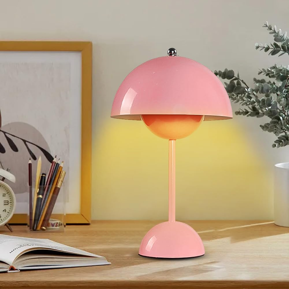 Rosa pilzförmige Tischlampe auf einem Holztisch. Ein Bild und eine Pflanze befinden sich im Hintergrund. Gelbes Licht.