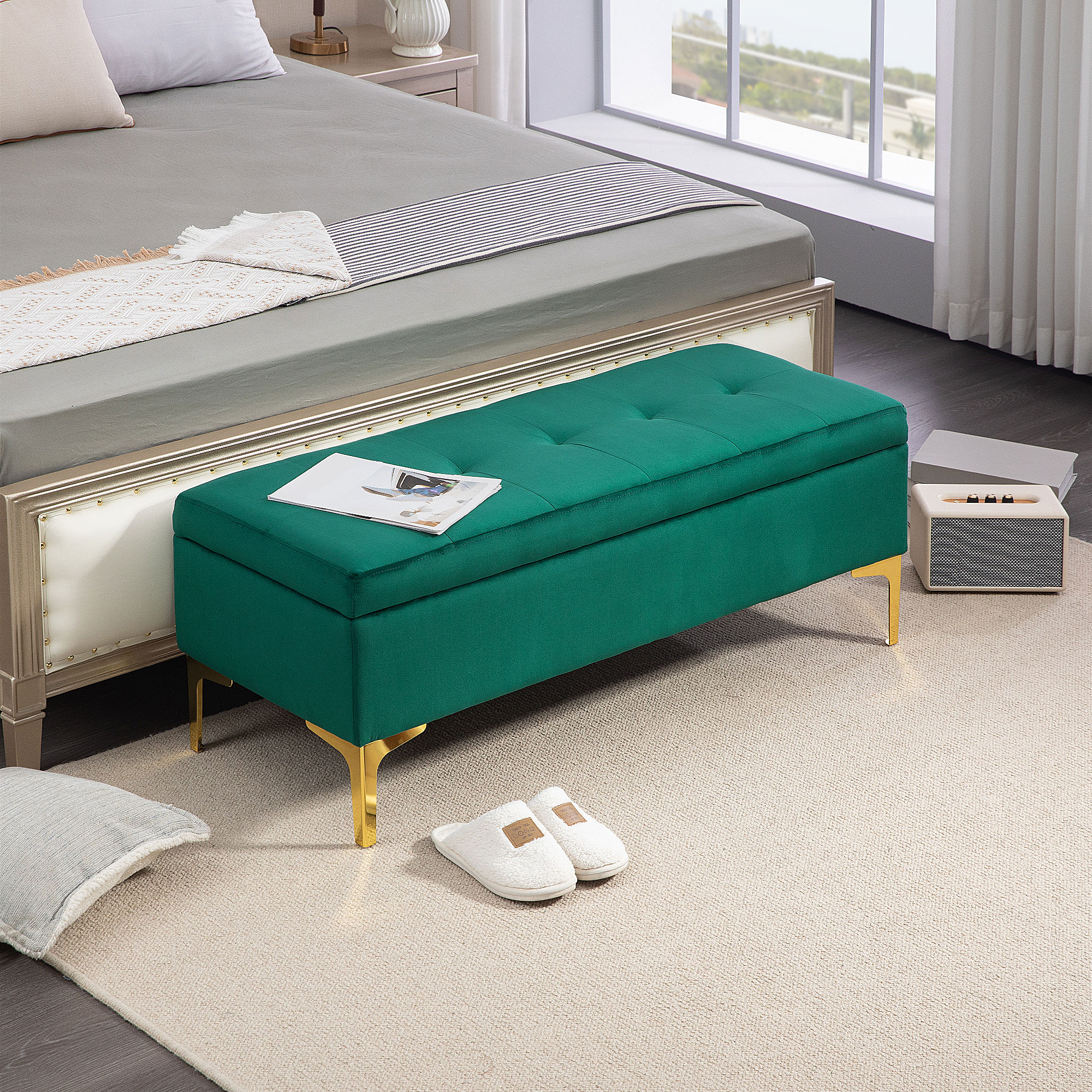 Eine grün gepolsterte Sitzbank mit goldenen Beinen, ein Bett im Hintergrund und Hausschuhe.