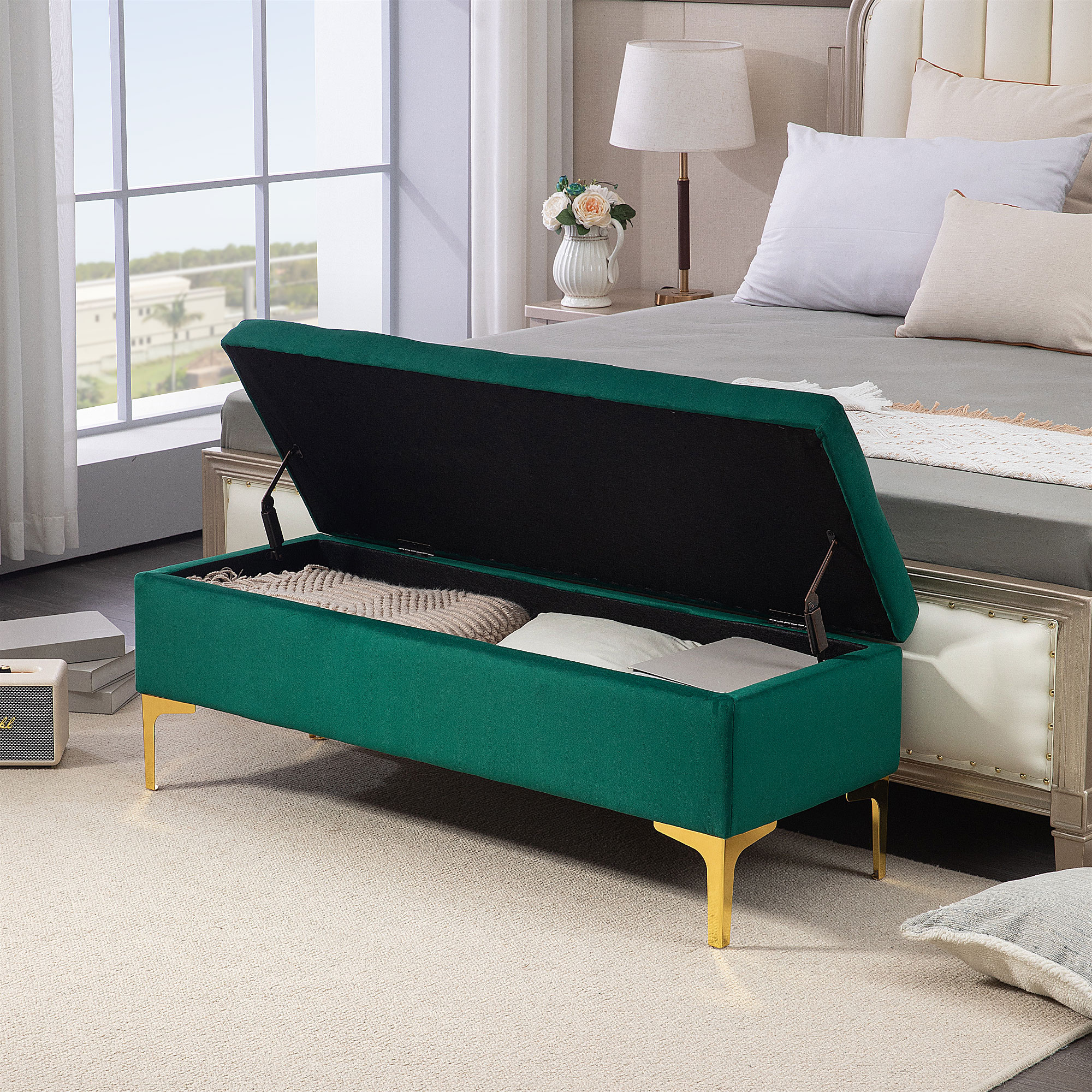 Eine grüne Aufbewahrungsbank mit offenem Deckel, Kissen und Dekoration. Goldbeine auf einem Teppich, ein Bett im Hintergrund.