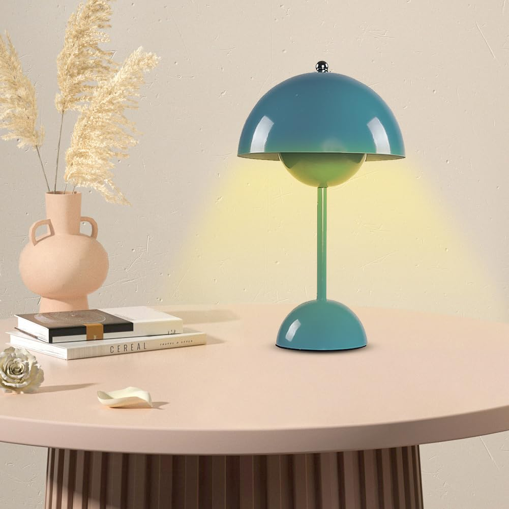 Eine blaue Tischlampe auf einem rosa Tisch. Eine Vase mit hohem Gras und Bücher sind in der Nähe. Die Lampe leuchtet.