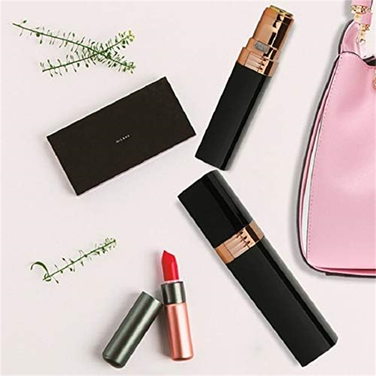 Kosmetika auf Weiß: schwarzes und goldenes Gerät, Lippenstift, rosa Handtasche, grüne Zweige und eine dunkle Schachtel.