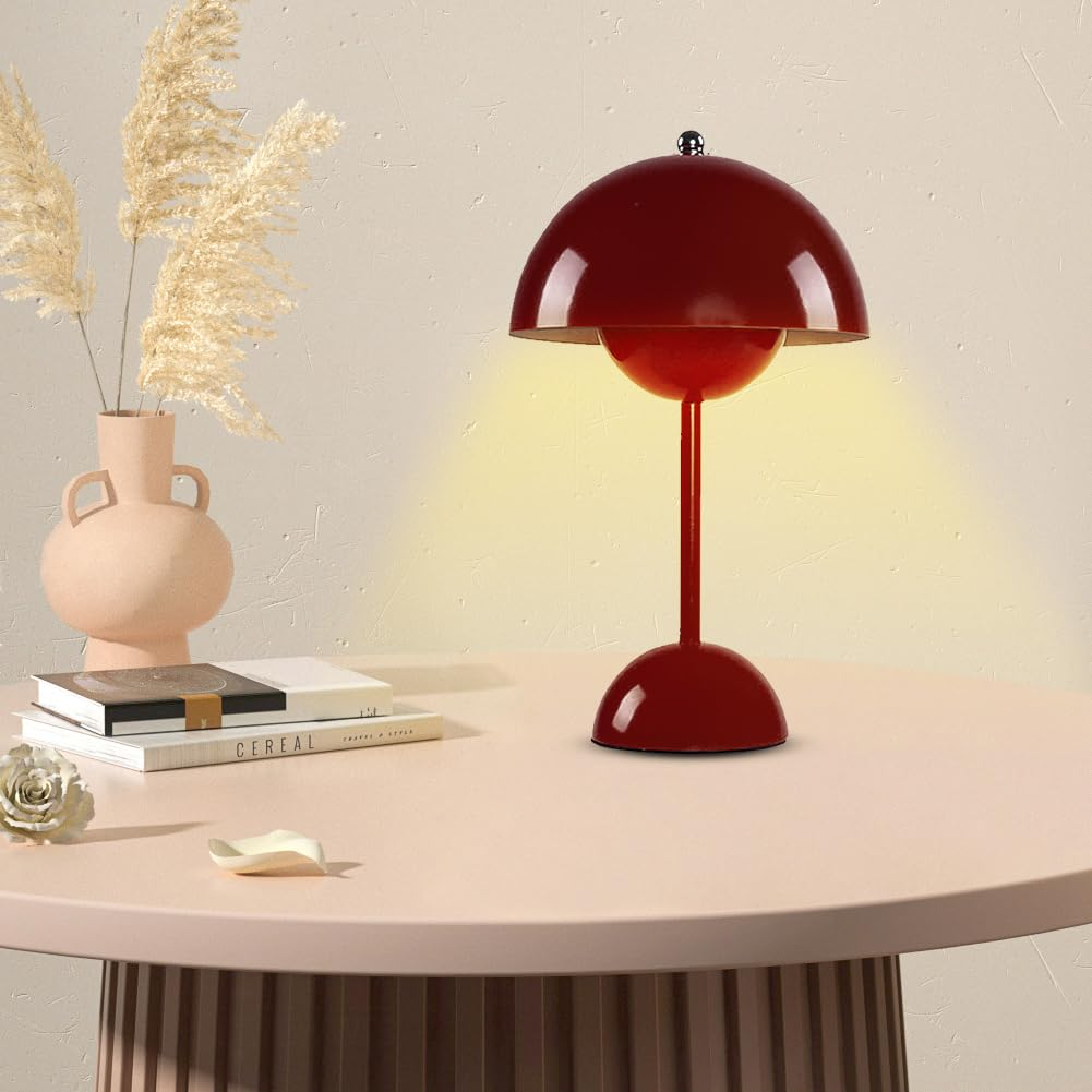 Eine rote Tischlampe auf einem Tisch. Eine rosa Vase, Bücher und eine weiße Schale stehen neben der Lampe.