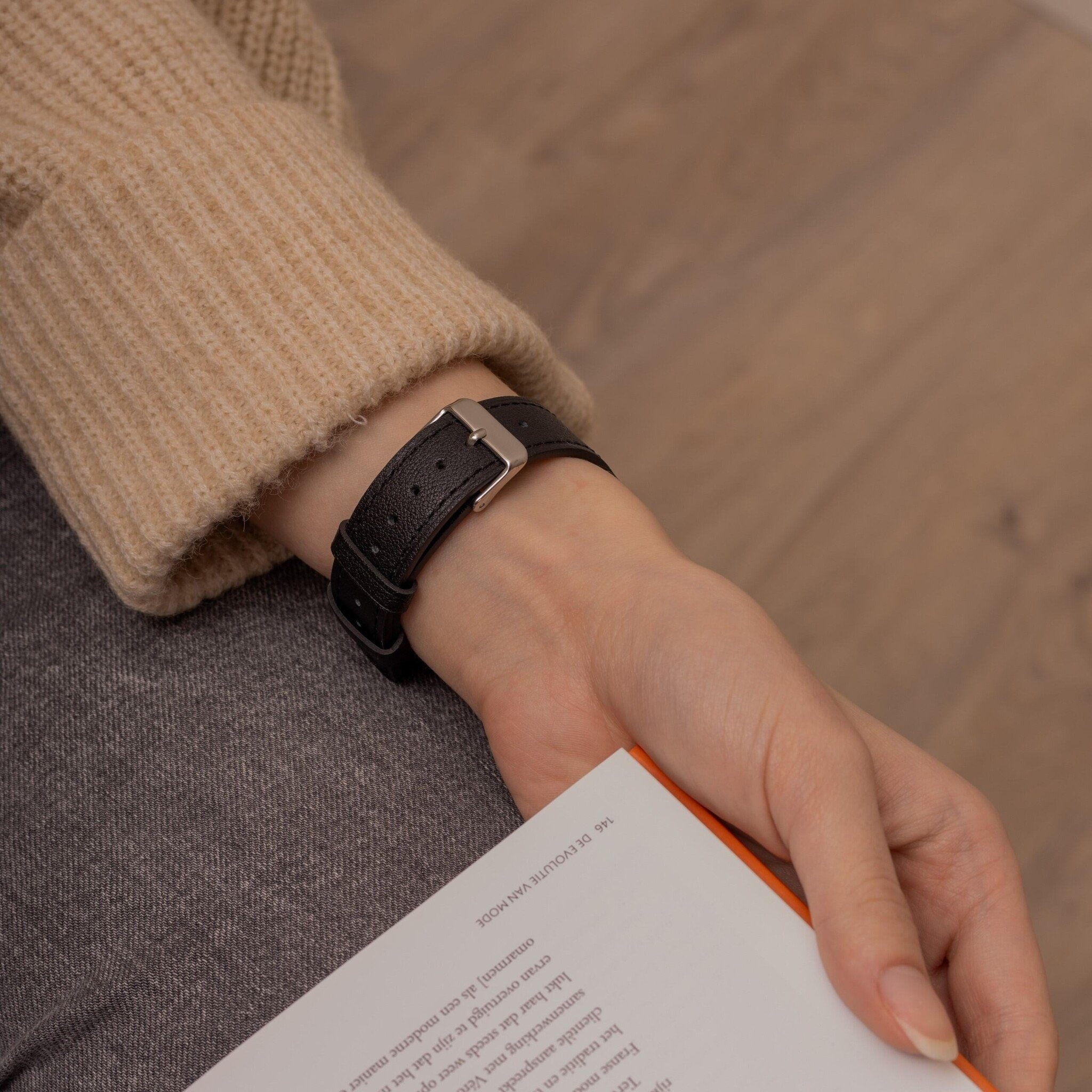 Handgelenk mit schwarzer Lederuhr, beigem Pullover, grauer Hose und Buch. Heller Holzboden im Hintergrund.